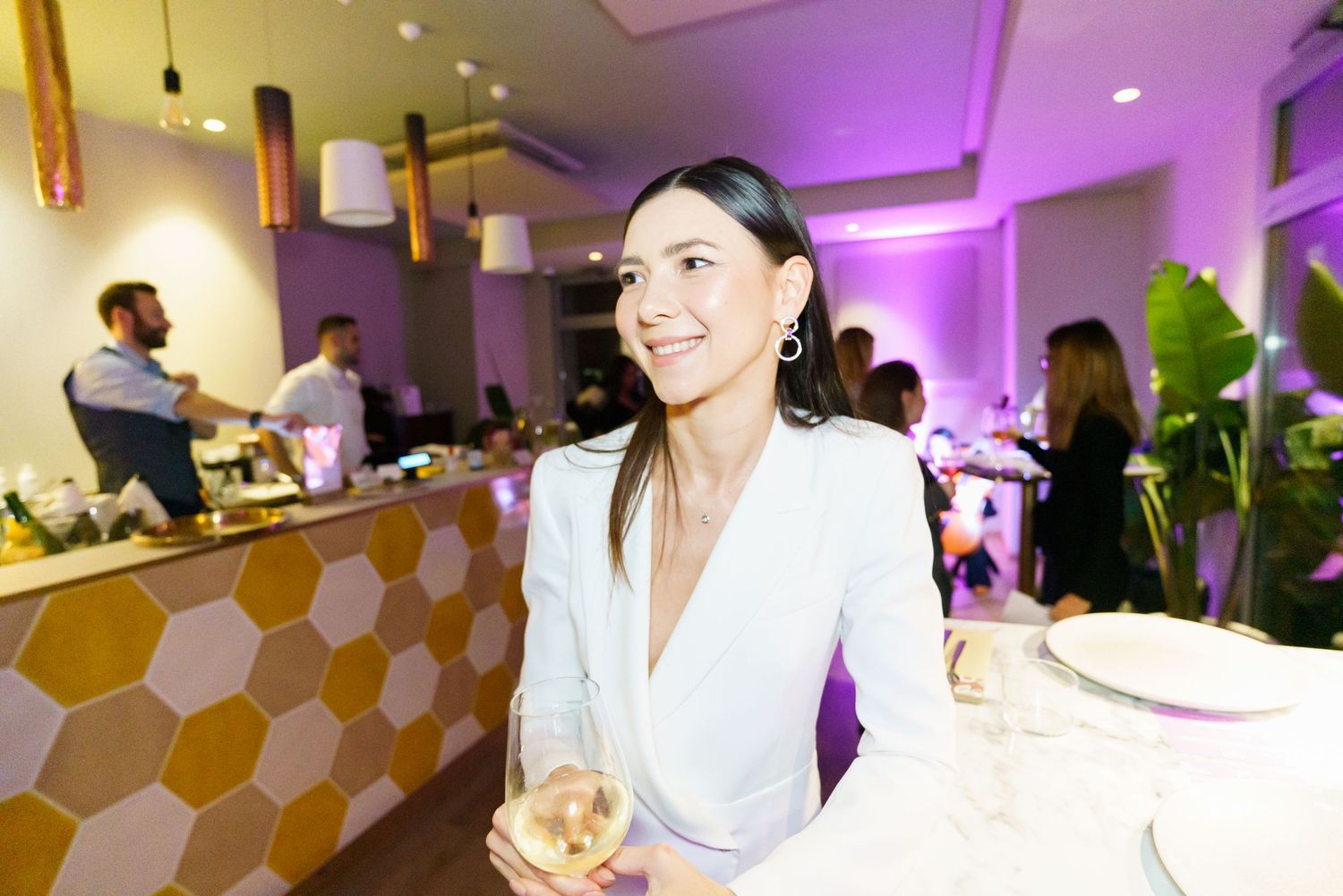 Elegant woman in white blazer holding a drink at a stylish restaurant with purple lighting.