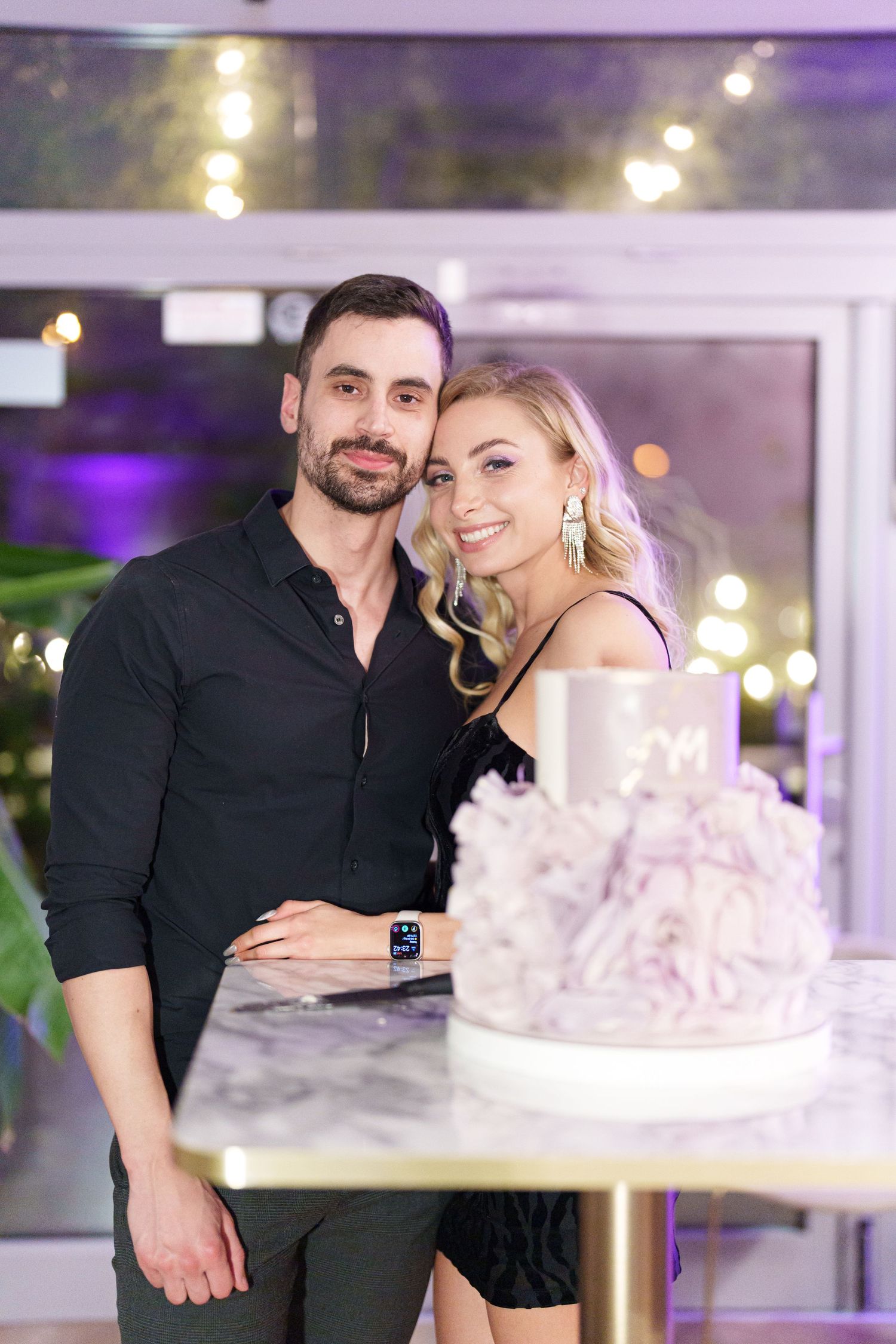 Smiling couple in black outfits posing together beside an elegant white tiered cake at a celebratory event.