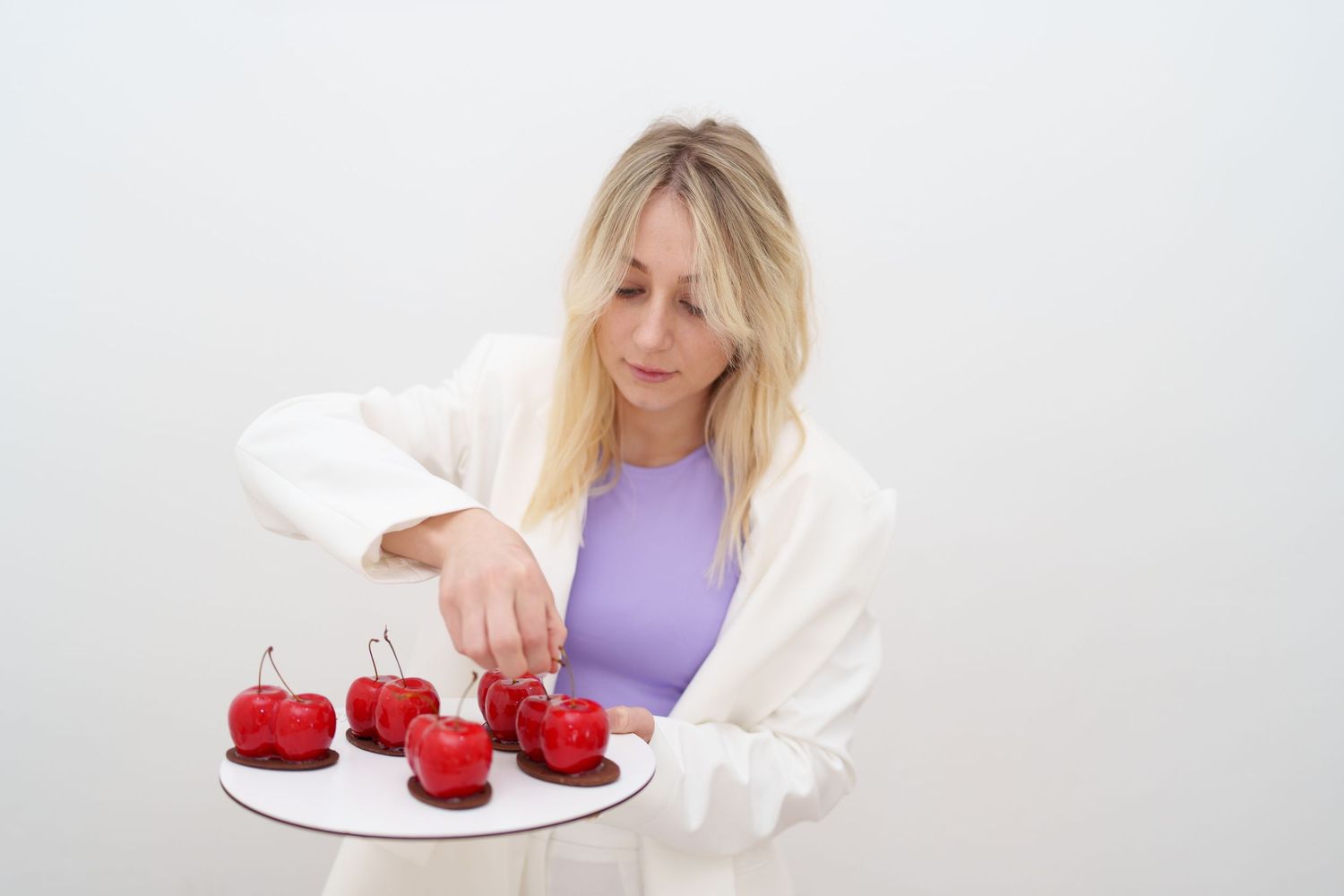 Blonde woman in white blazer arranging red cherry-shaped chocolates on a white plate against light background.