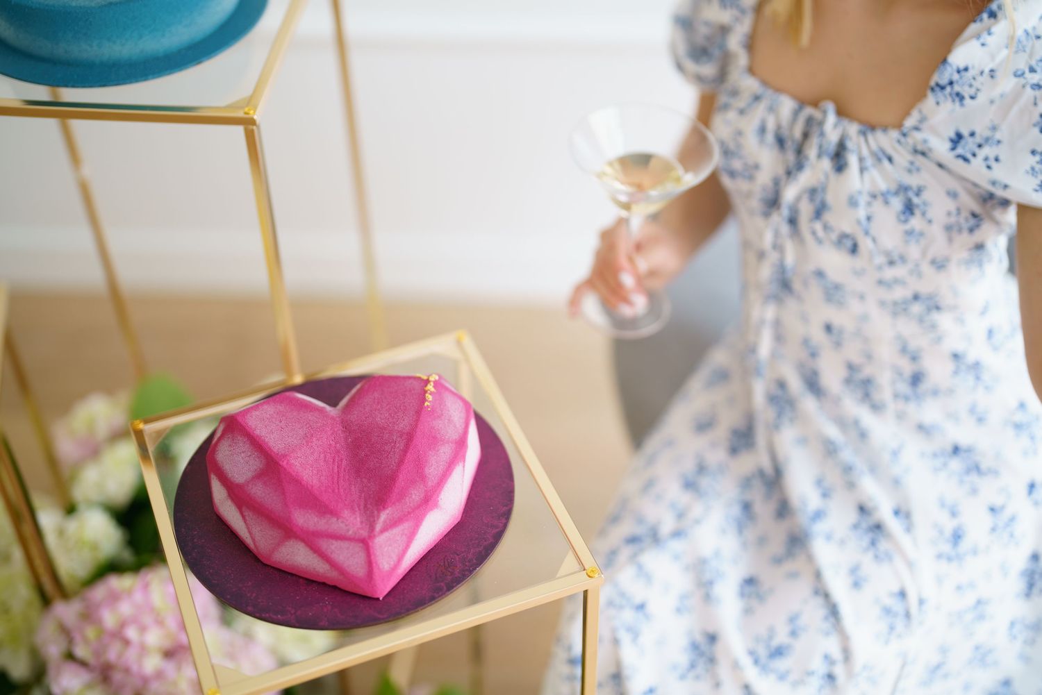 A pink geometric heart-shaped cake on a gold stand beside a woman in a floral dress holding a champagne glass.