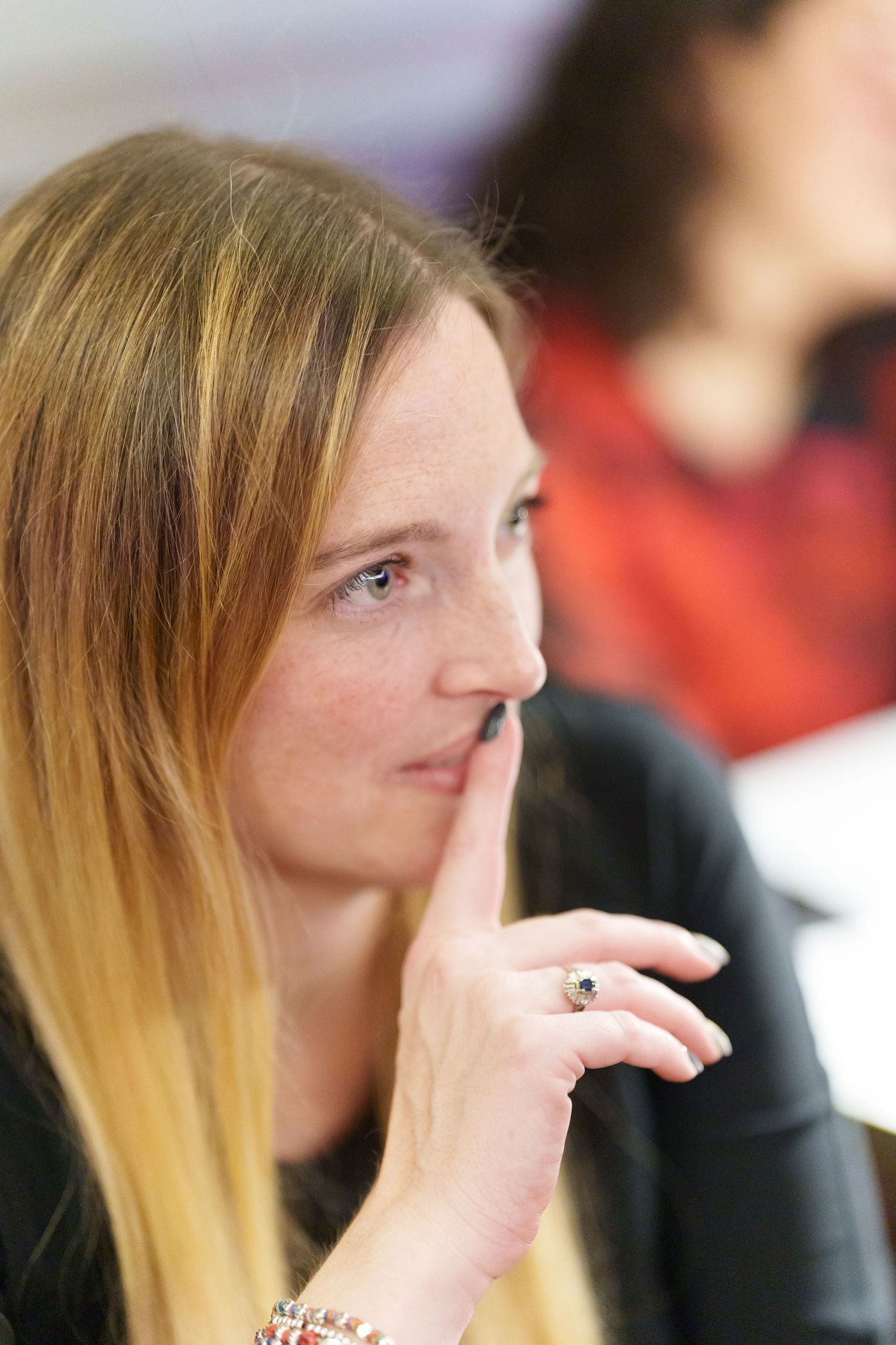 Woman with long blonde hair holding finger to lips in a shushing gesture at an indoor event.