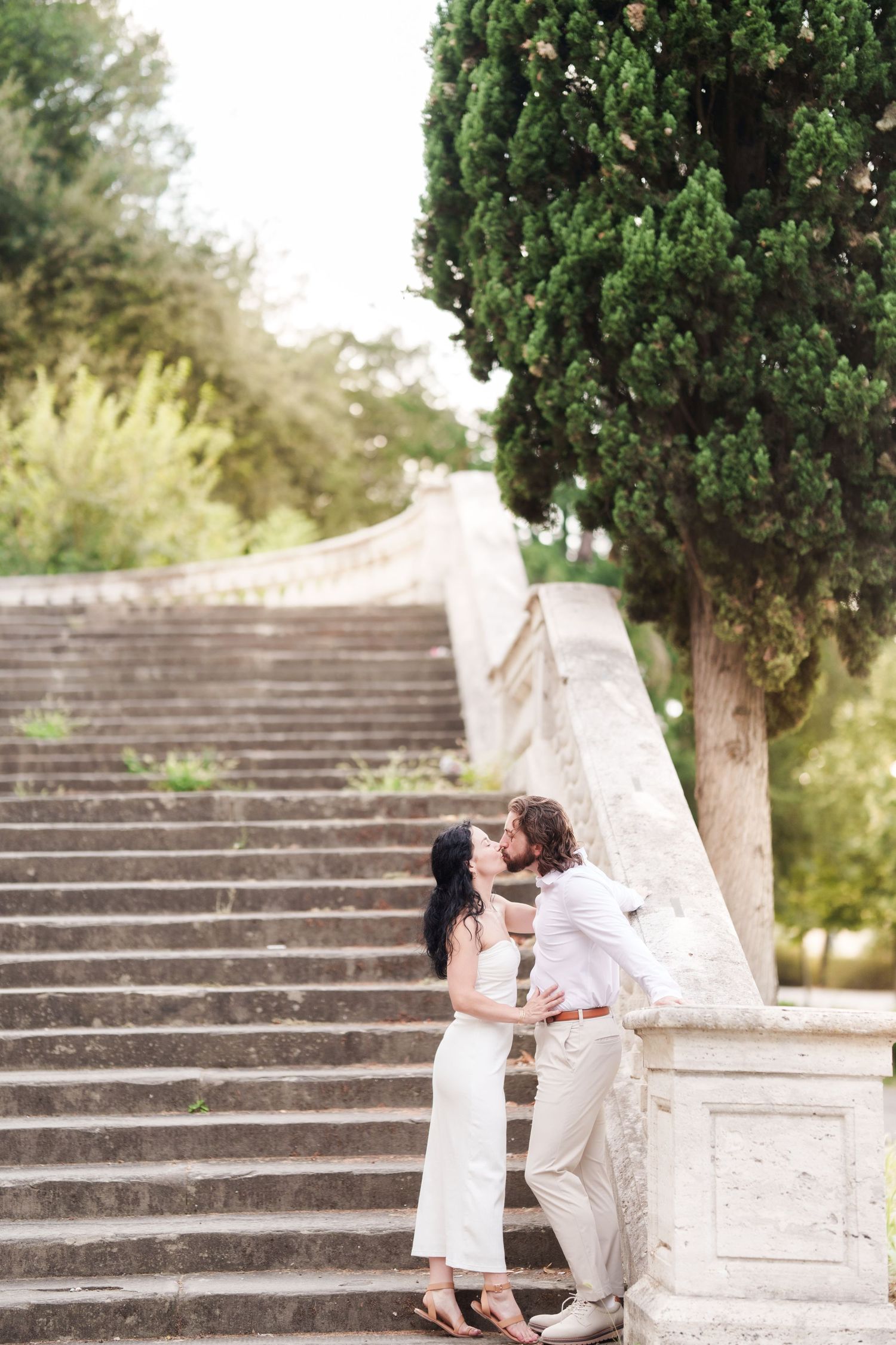 A romantic couple shares a tender moment on elegant stone steps surrounded by lush greenery in a garden setting.