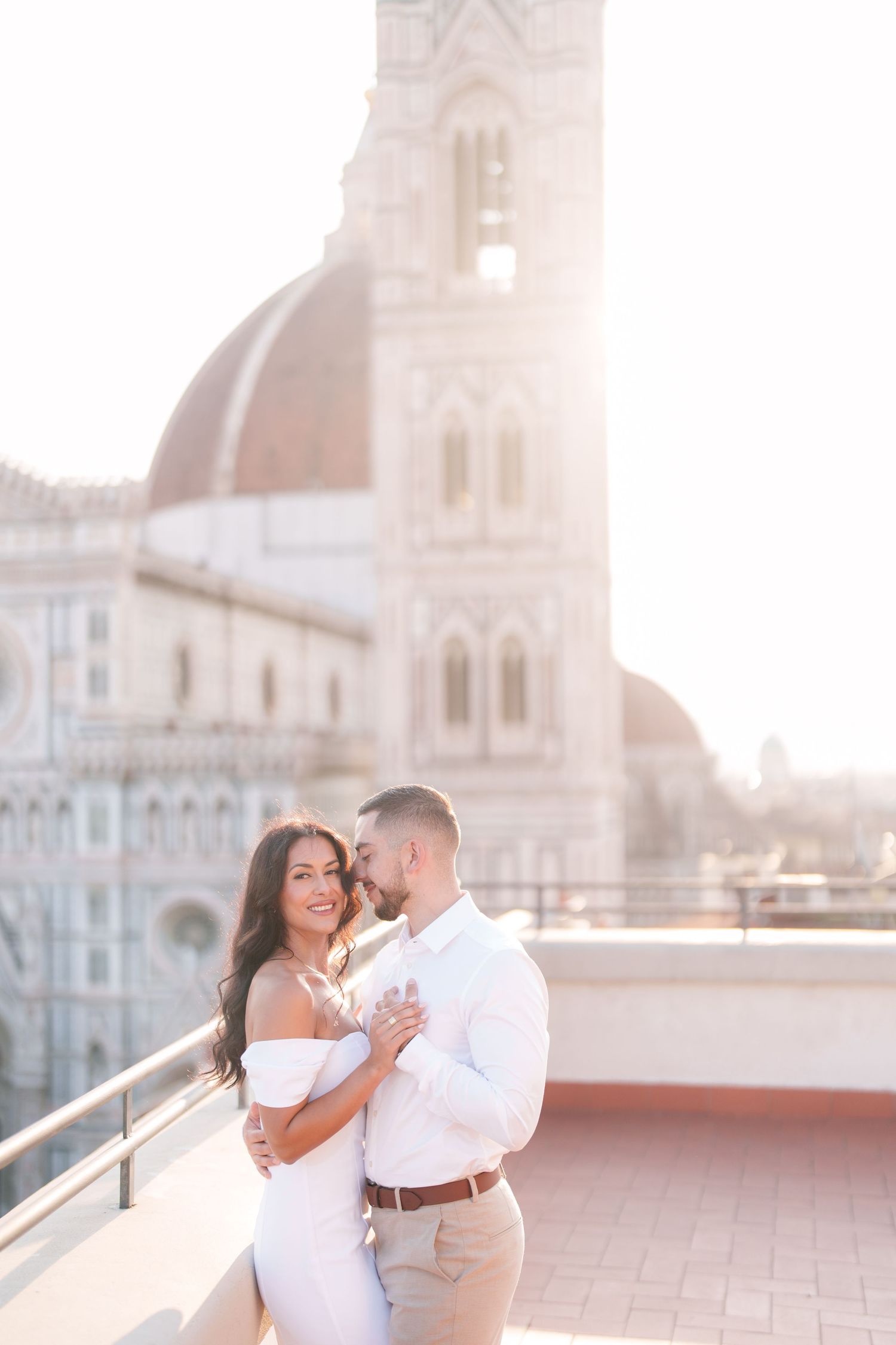 A couple in white outfits shares romantic moments on a rooftop terrace with a European cityscape in the background.