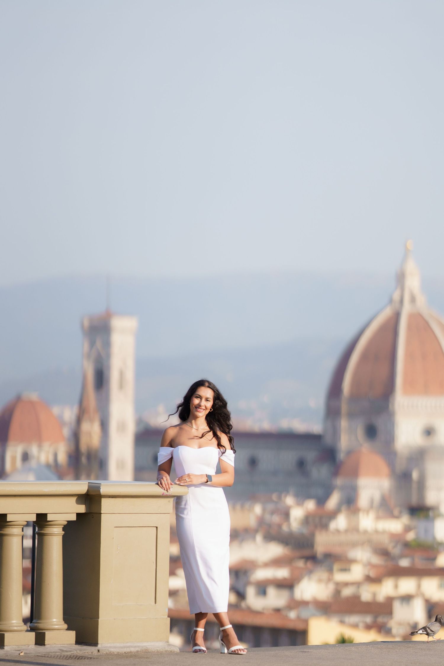 A person in a white dress poses against the iconic backdrop of Florence's Duomo cathedral and cityscape on a sunny day.