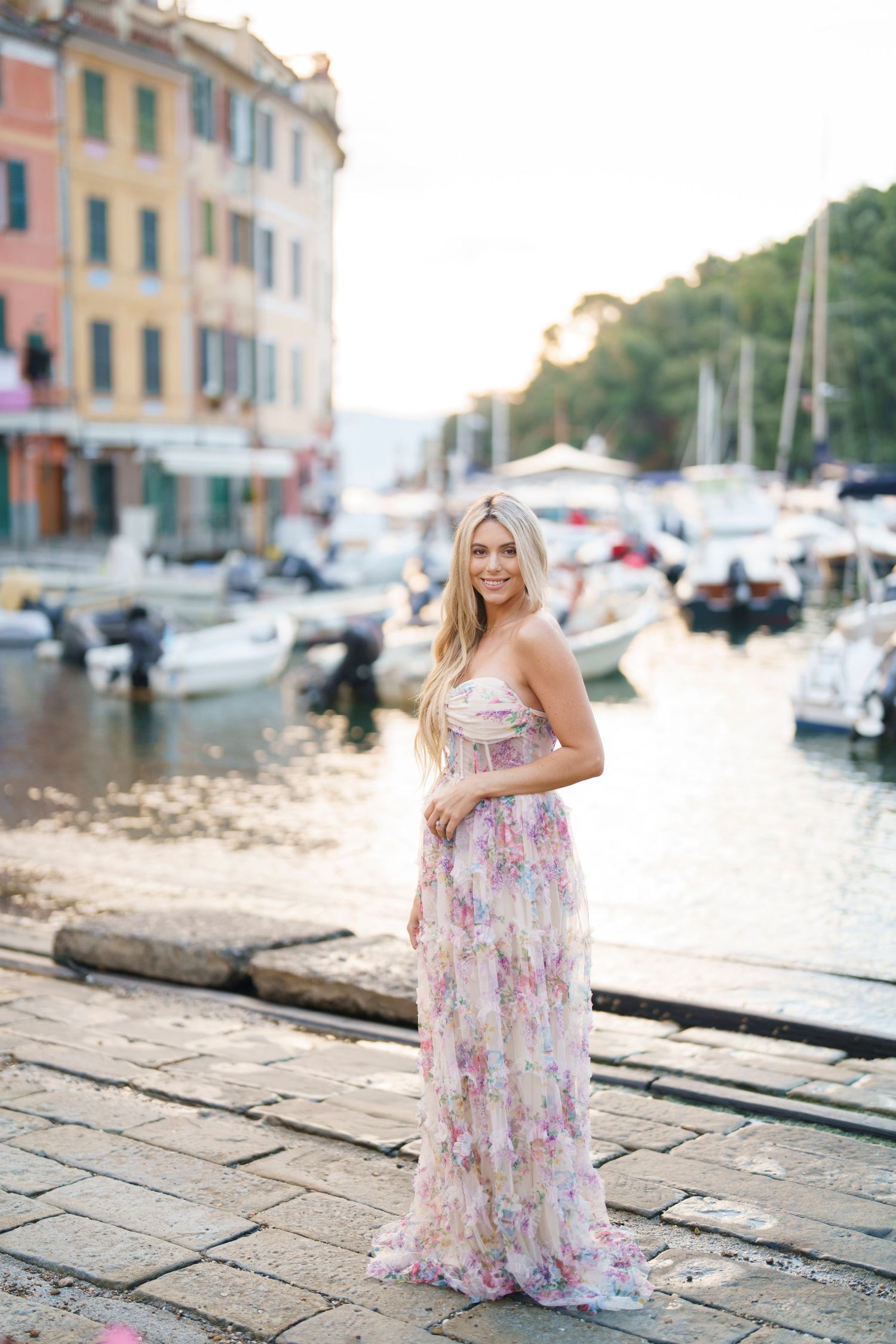 A person in a floral dress poses along a waterfront marina with boats and buildings in the background.