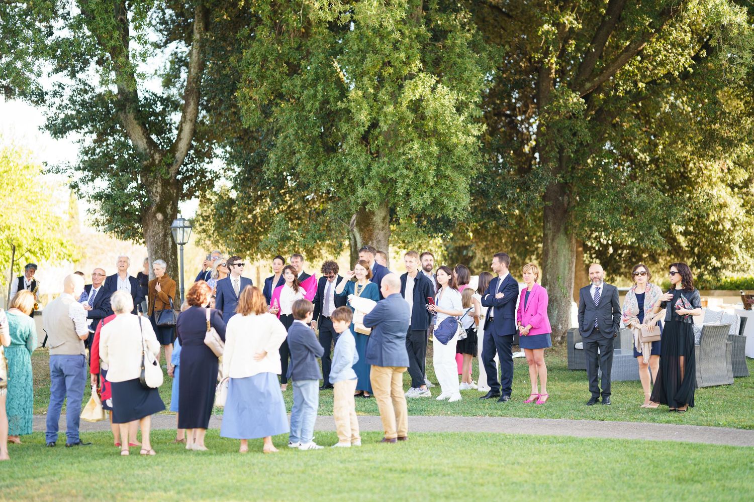 Large group of wedding guests gathered outdoors under green trees.