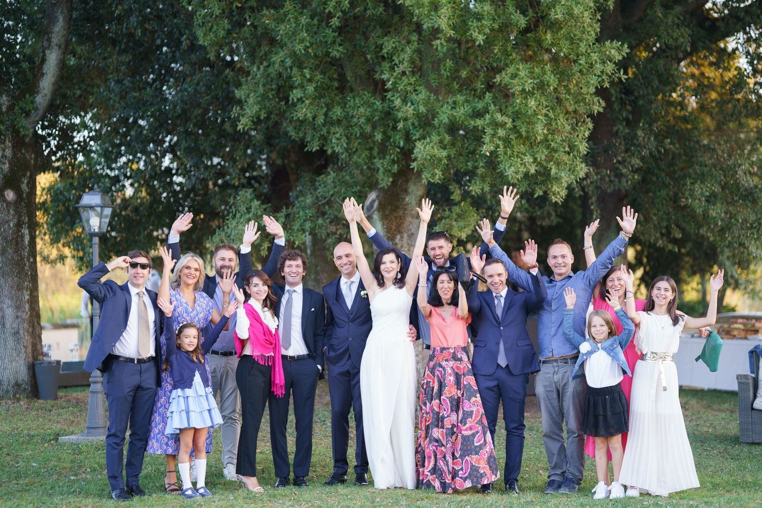 Wedding group celebrates with raised arms and cheers under trees at outdoor reception.