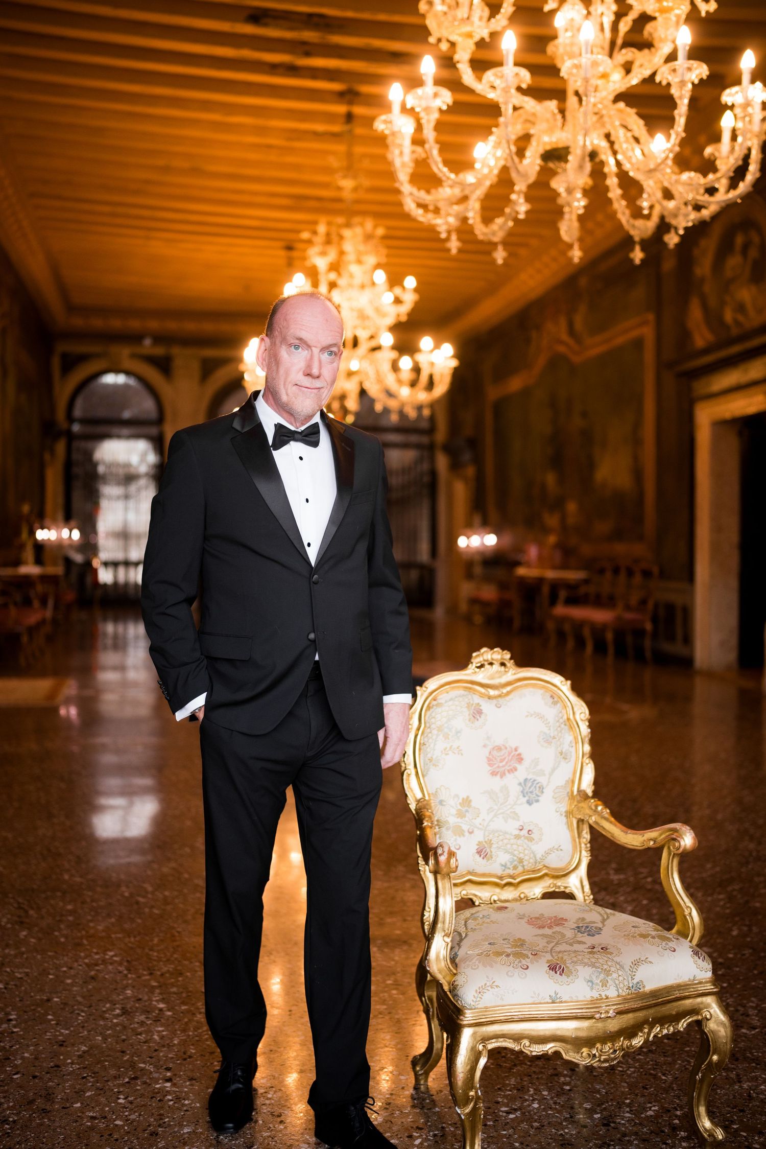 Elegant ballroom interior with ornate crystal chandeliers, gilded chair, and person in formal black tuxedo.
