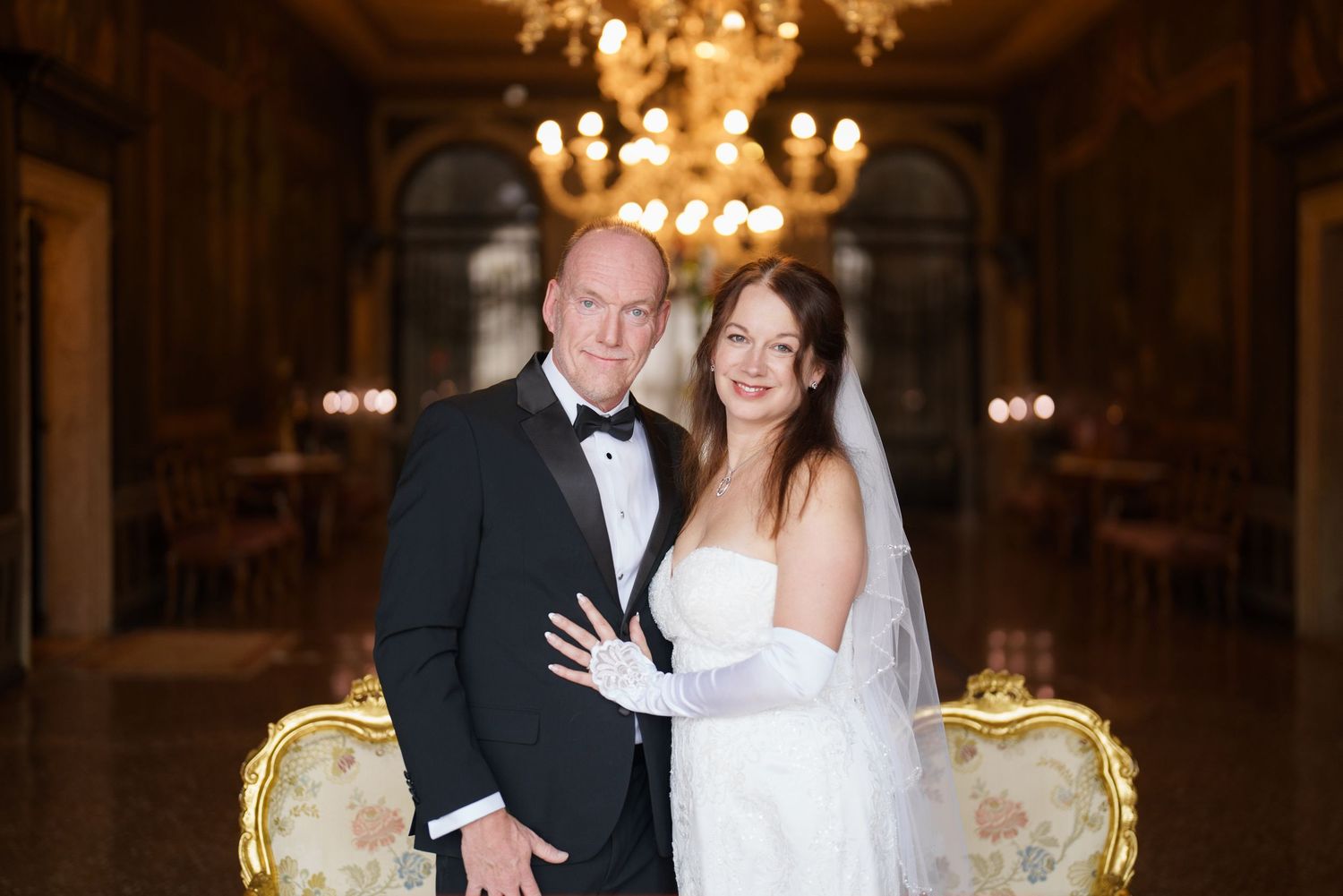 A bride and groom pose together in an elegant ballroom with ornate chandeliers and gilded furniture.