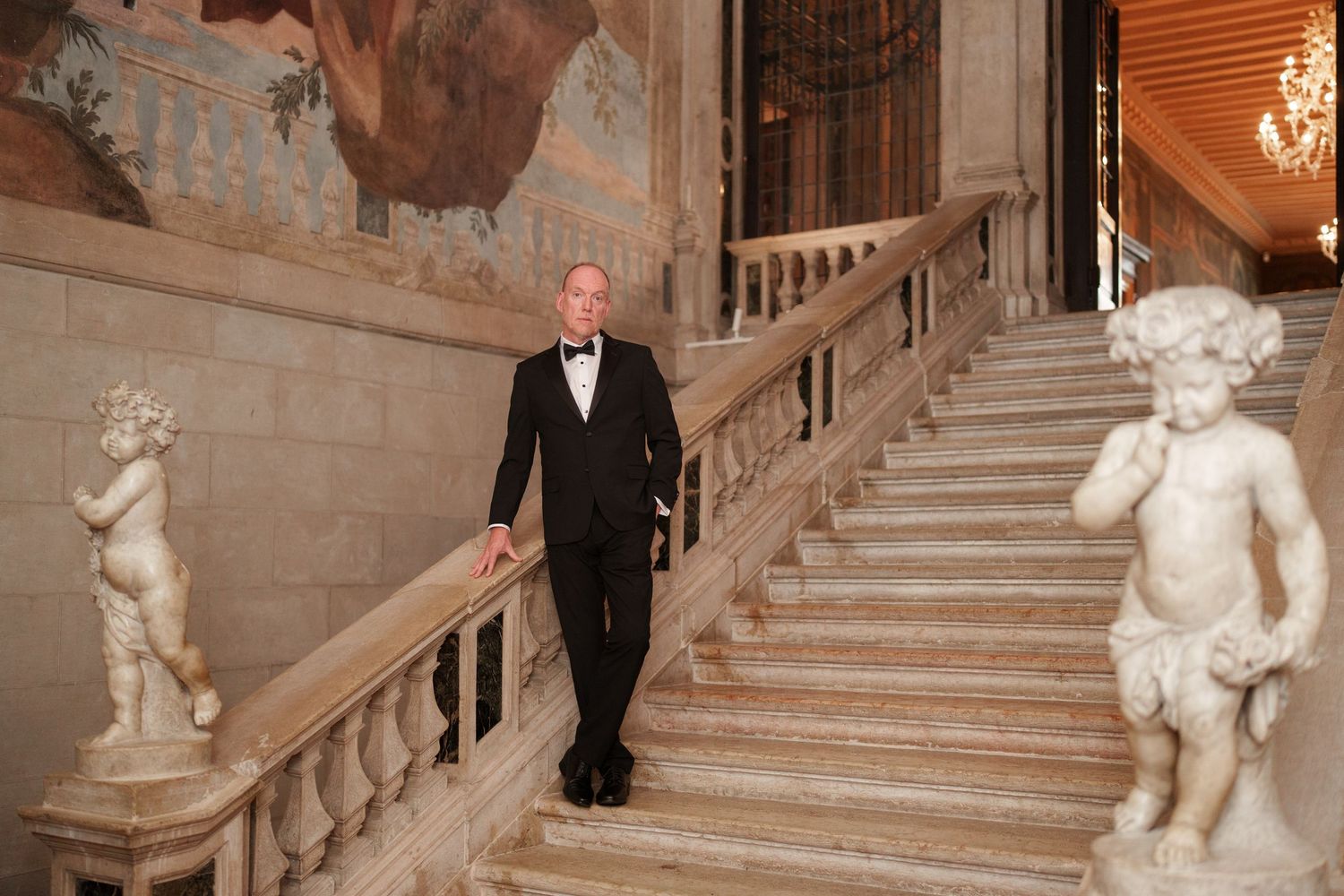 A person in formal attire descends an ornate marble staircase adorned with cherub statues and elegant railings.