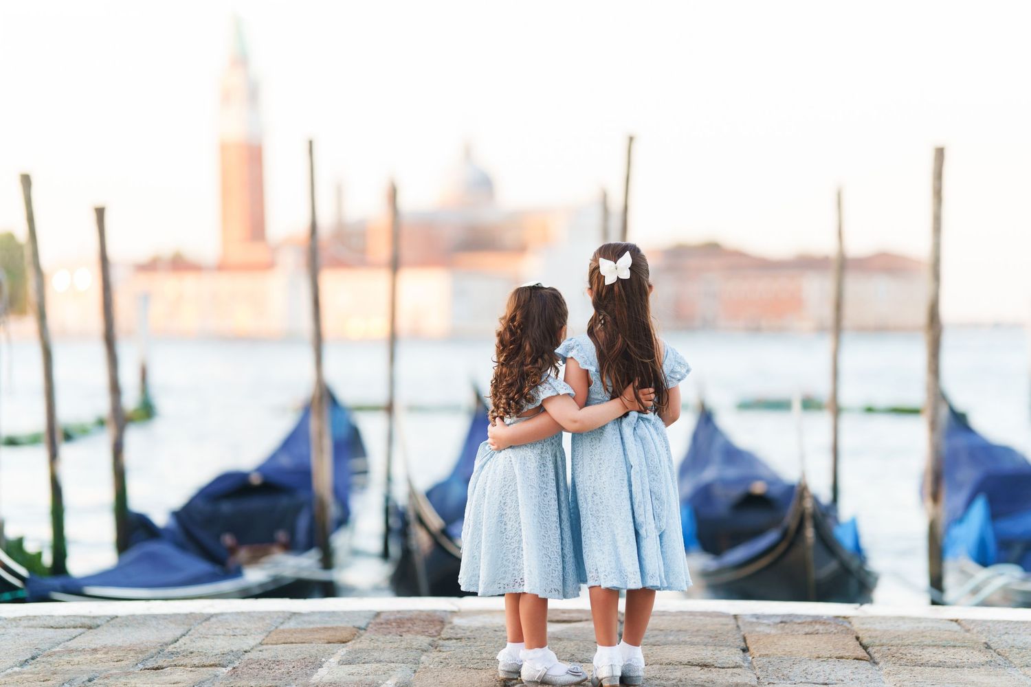 Two people holding drinks stand on a beach at sunset with gondolas and the Venice skyline in the background.