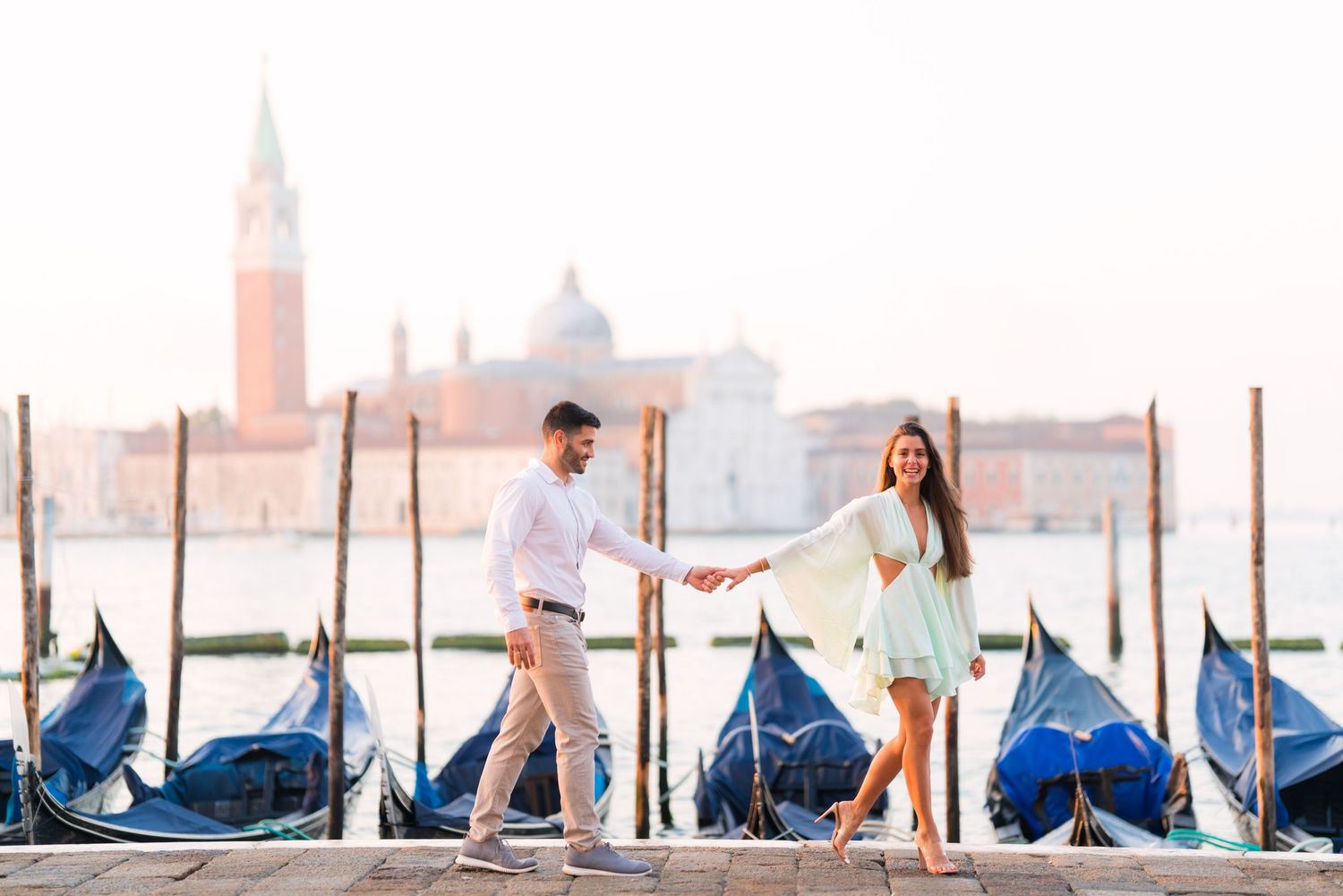 Row of traditional Venetian gondolas docked along wooden posts in the lagoon with historic buildings in background.