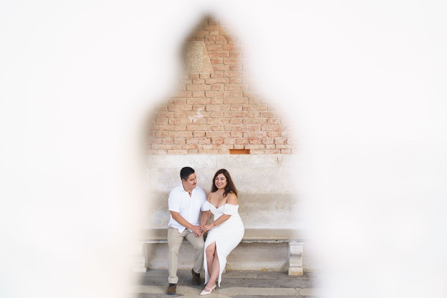 Couple in white outfits poses together against a textured brick wall in a series of romantic engagement photos.