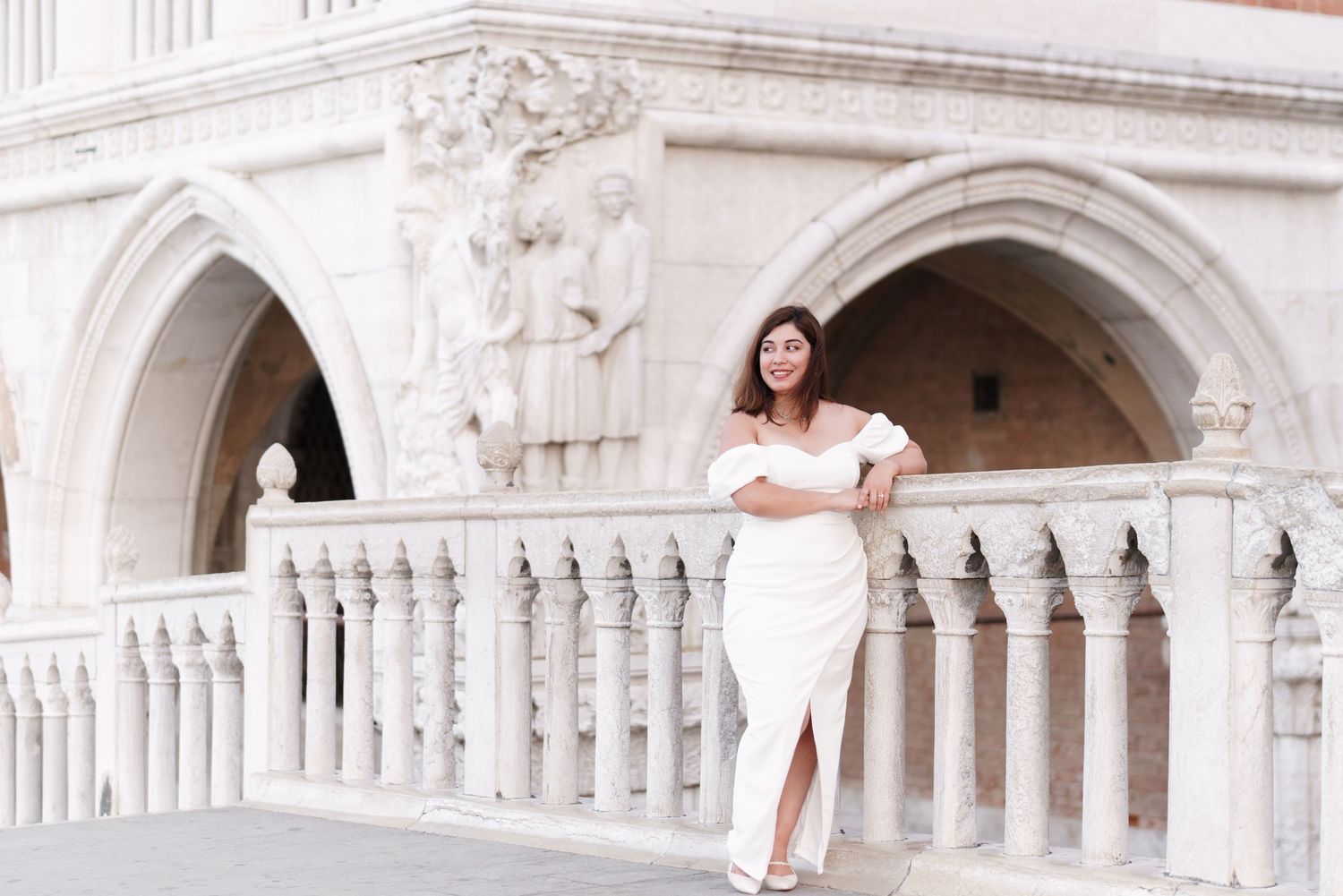 A couple in wedding attire poses on ornate white marble steps of a historic building with arched architecture.
