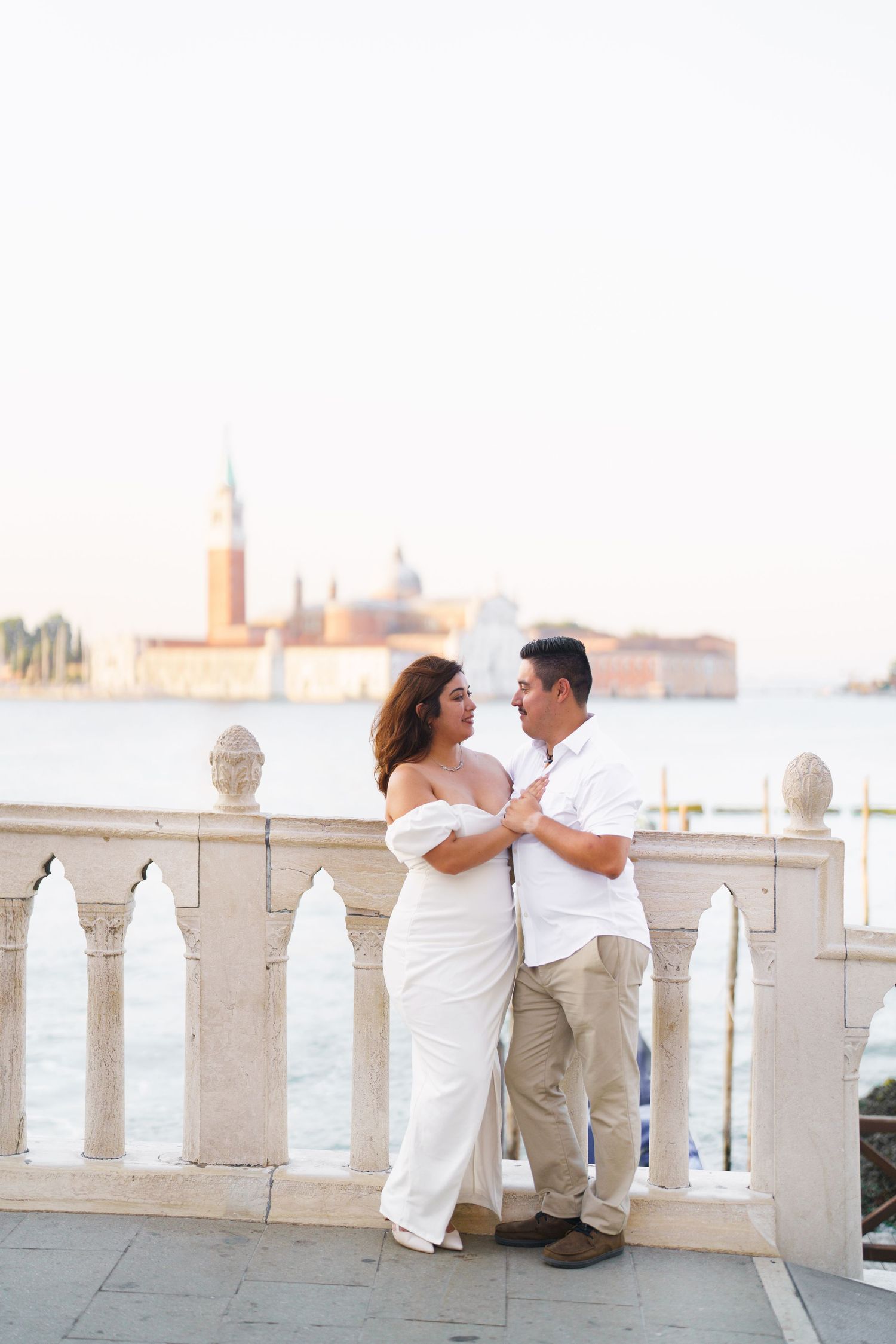 Couple in white outfits sitting on a balcony overlooking a scenic waterfront cityscape at sunset.