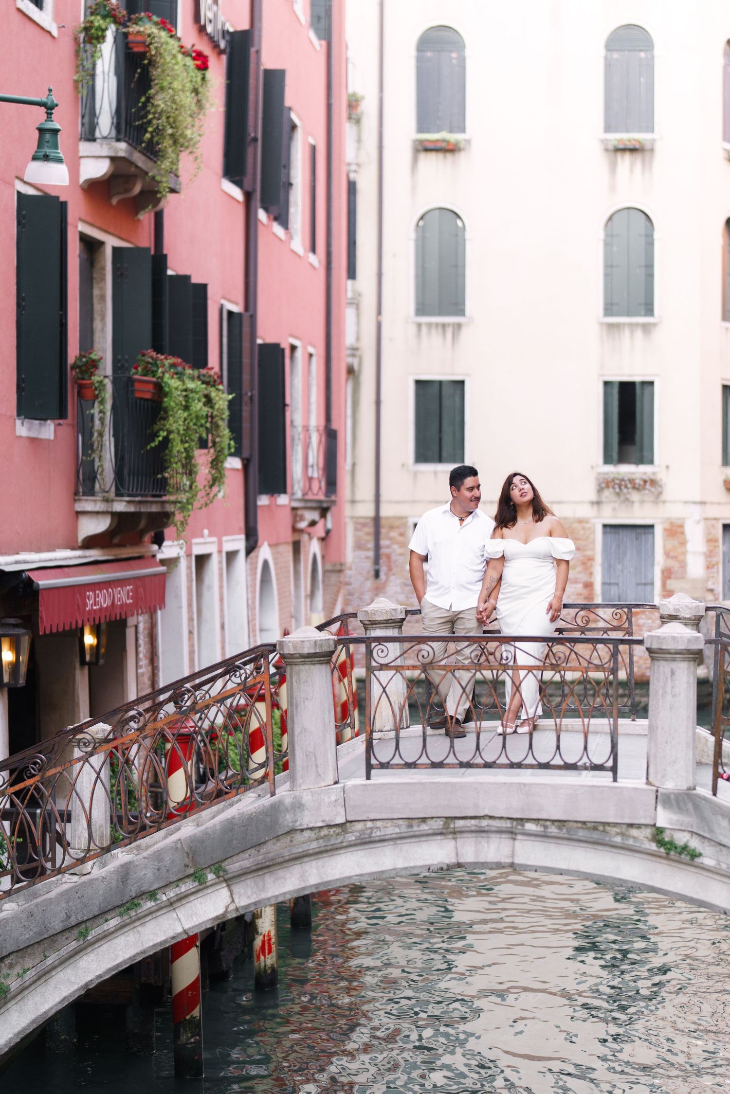 Couple walking across a picturesque arched bridge over a Venice canal with pink buildings in the background.