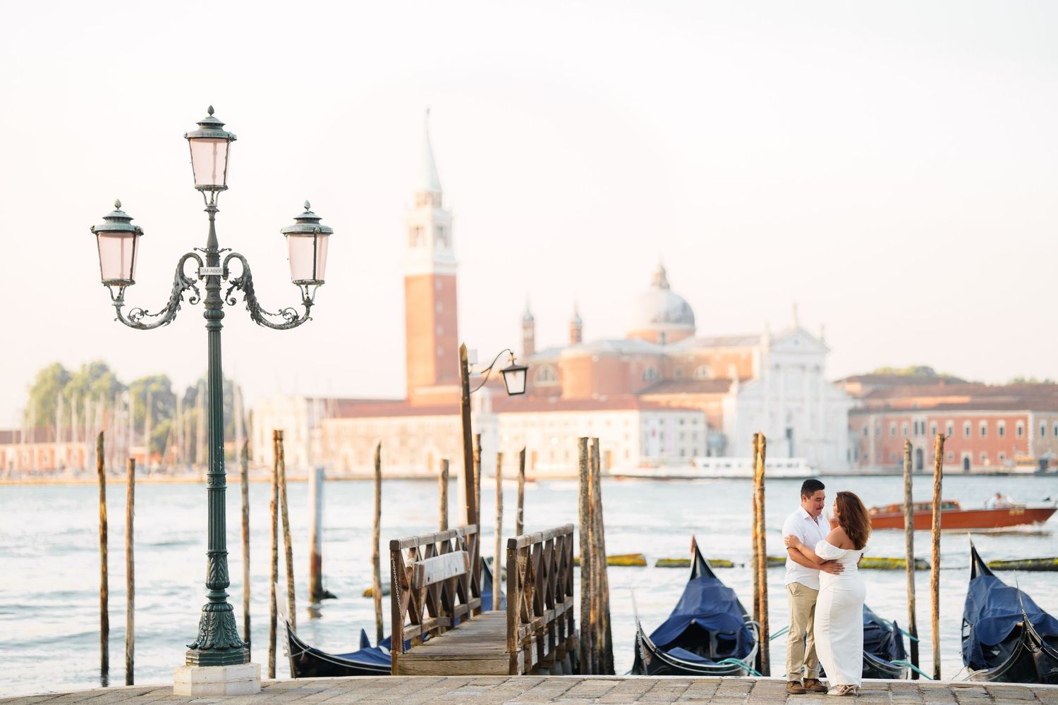 Romantic Venice waterfront scene with ornate lamp post, moored gondolas, and San Giorgio Maggiore church in the background.
