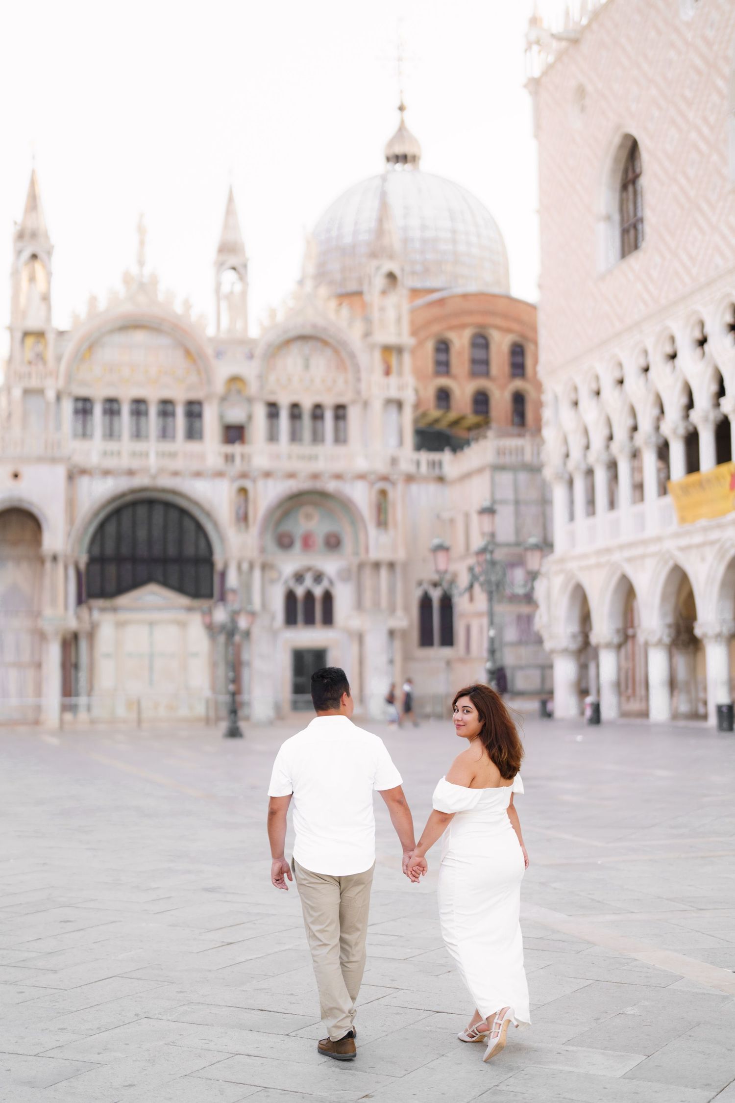 Couple walking hand in hand through St Mark's Square in Venice with the basilica's ornate architecture in background.