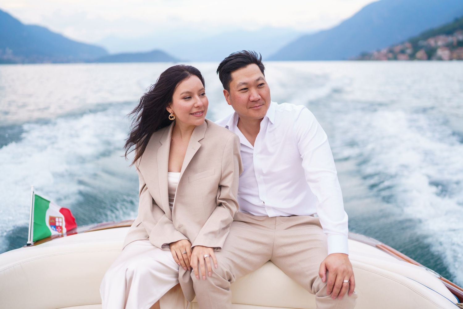 A couple in light-colored outfits share romantic moments together on a boat and beach with mountains in the background.