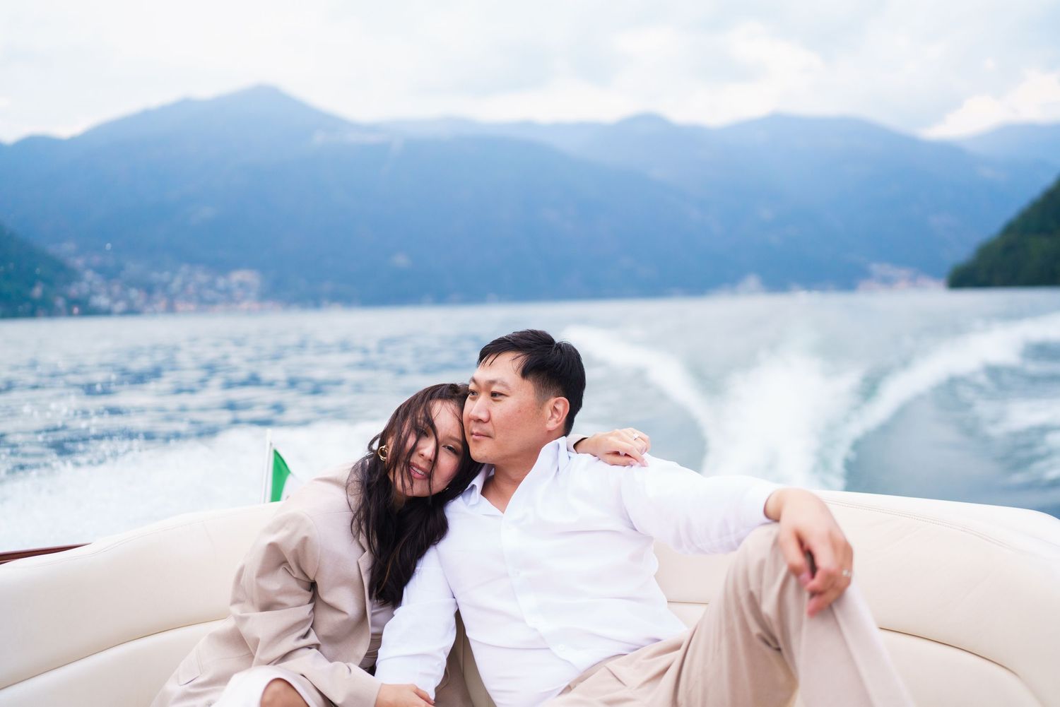 Romantic couple shares intimate moments by the water with mountains and cloudy skies in the background.