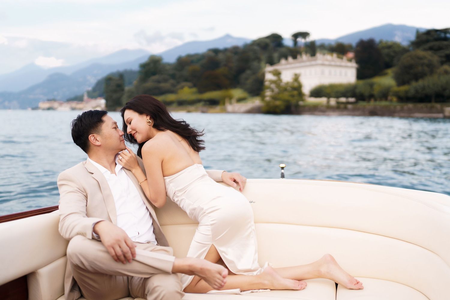 A couple in light-colored attire share an intimate moment on a luxury boat with a coastal villa and mountains in the background.