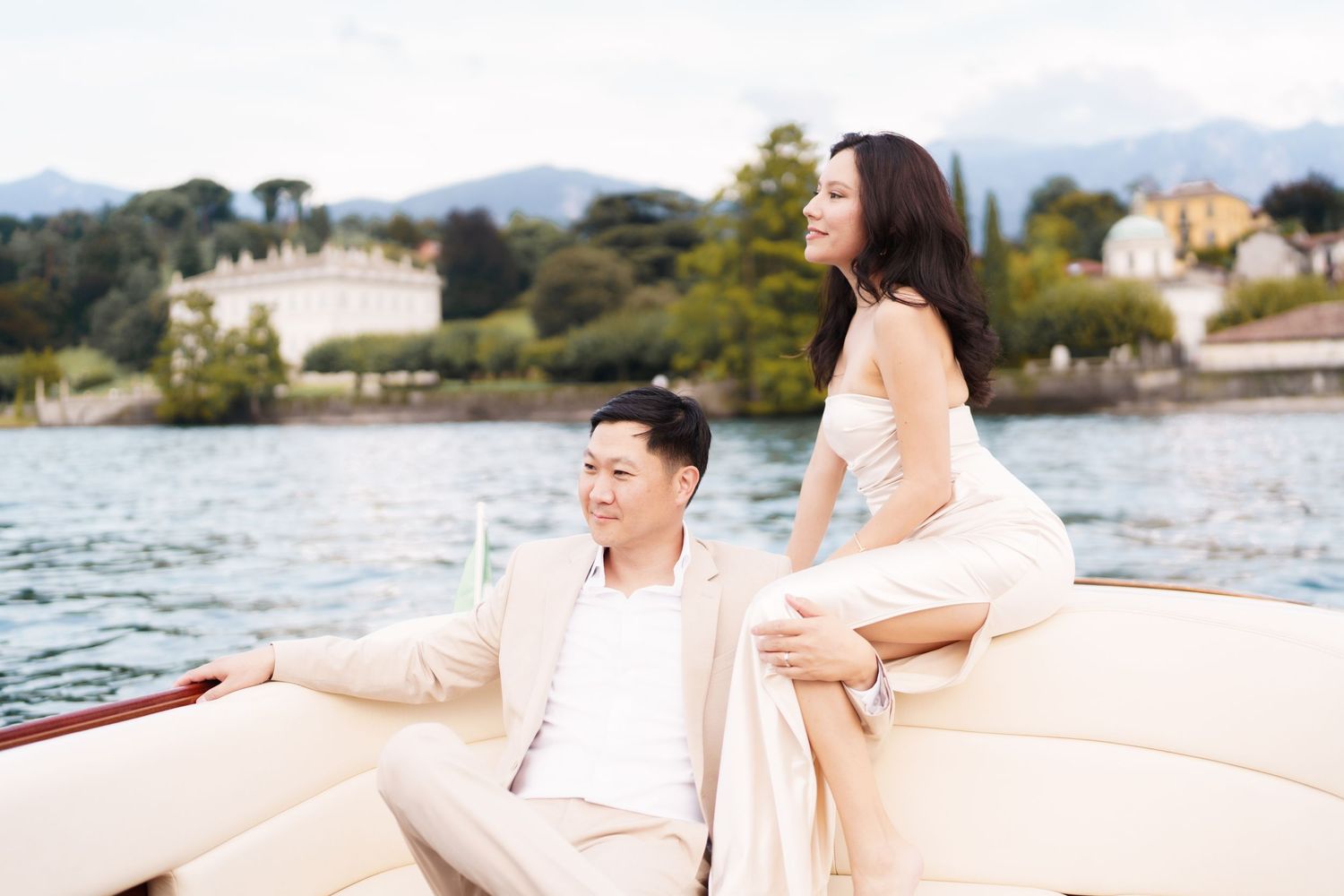 Couple in white outfits relaxing on a boat deck by a scenic waterfront with mountains in the background.