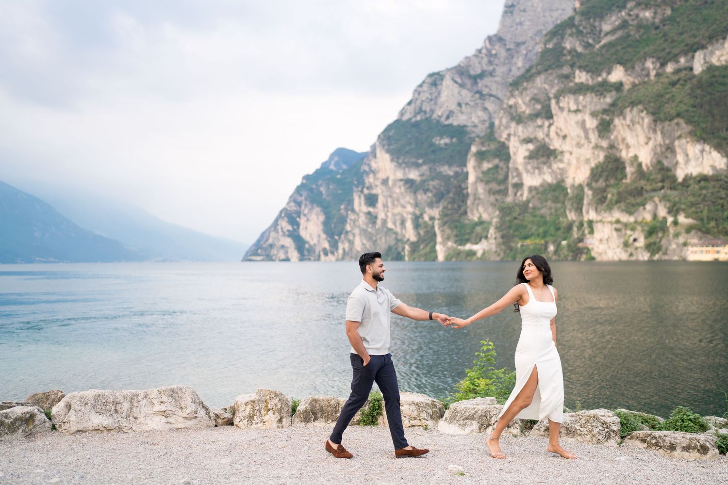 A romantic couple walks hand in hand along the scenic shores of Lake Garda in Italy with towering cliffs in the background.
