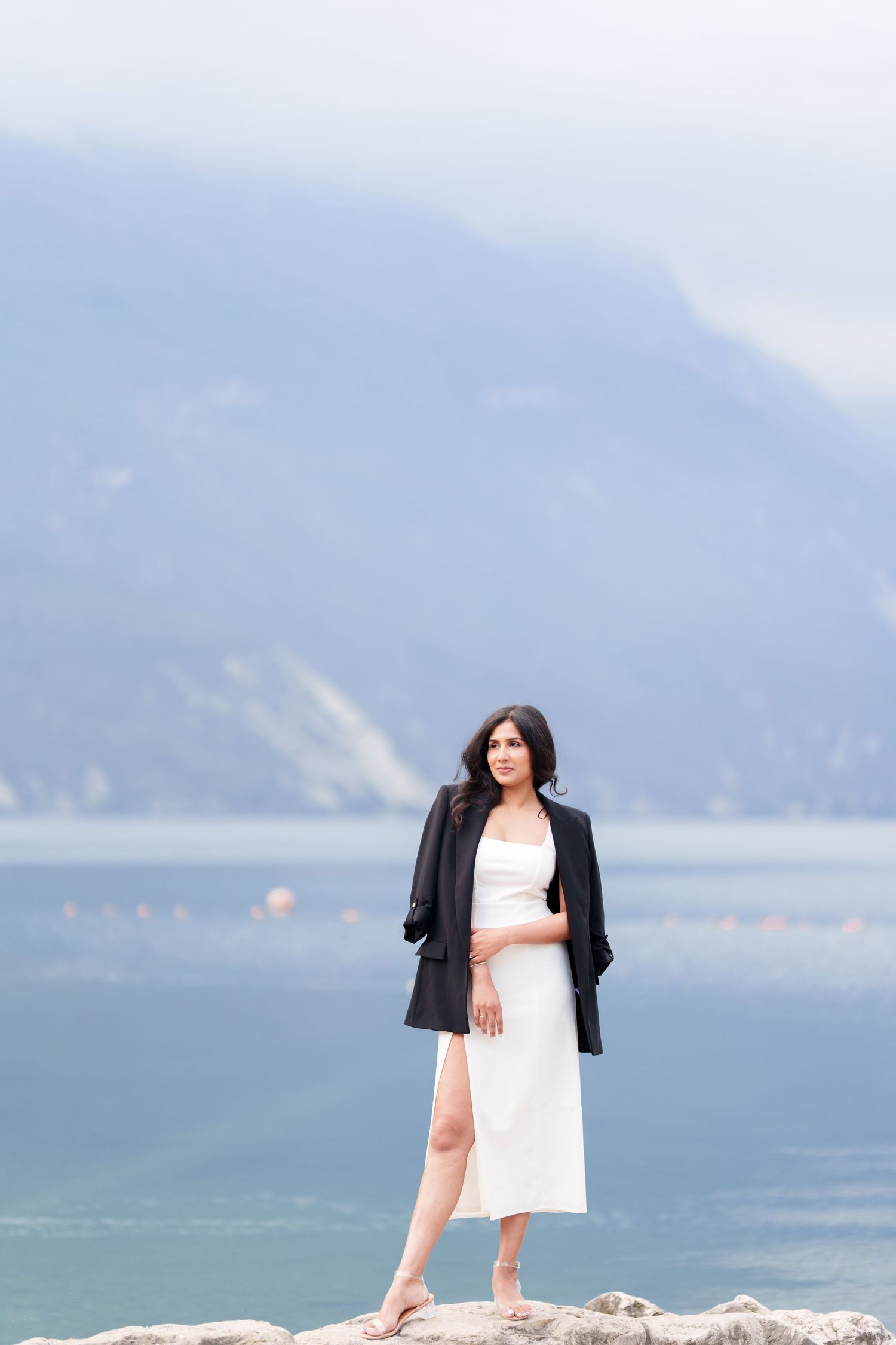 Person in white dress and black jacket poses on rocky shore with misty mountains and lake in background.