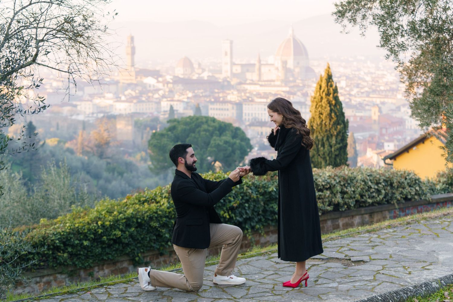 A romantic proposal moment on a hilltop overlooking a misty cityscape with cypress trees and a stone path.