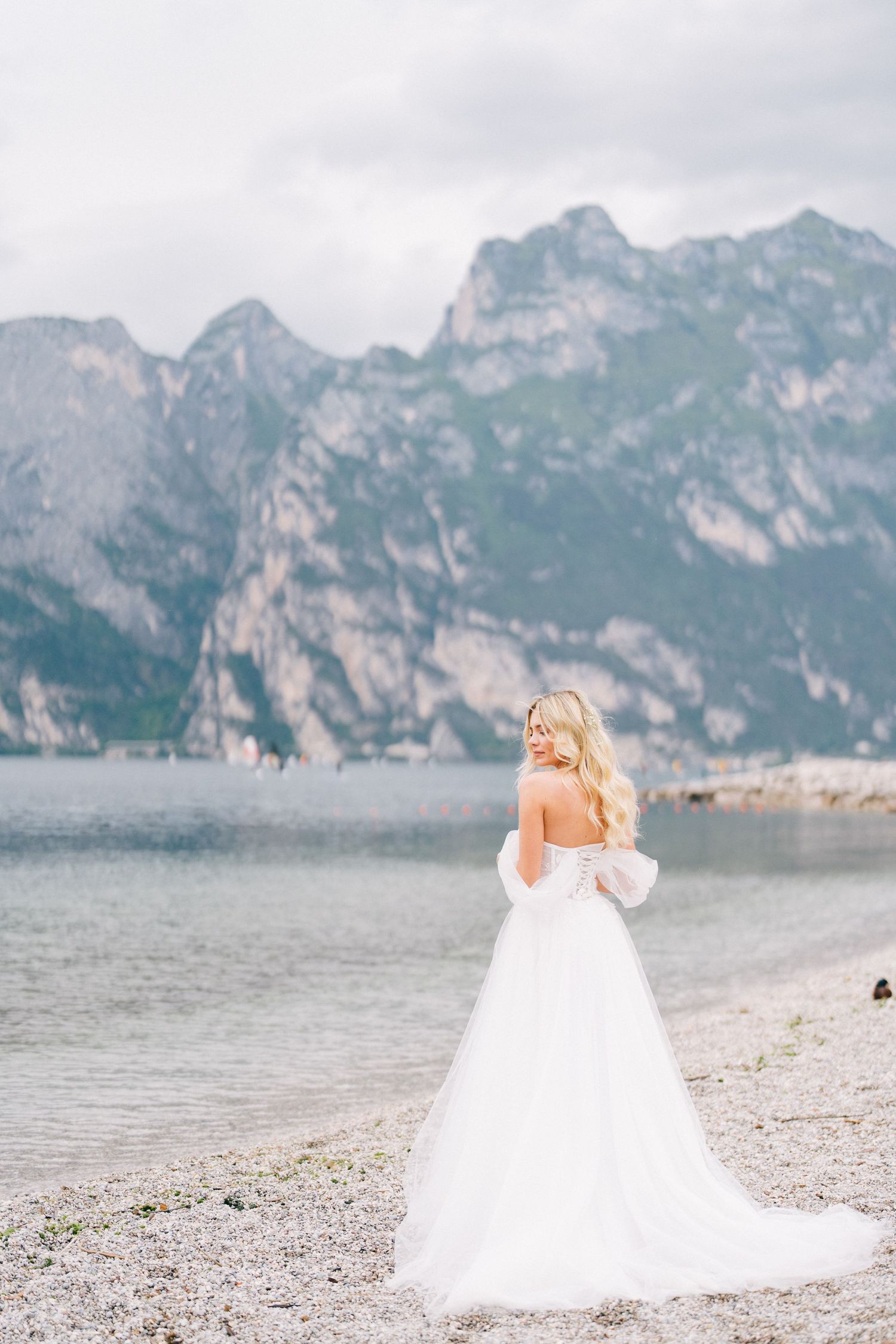 A bride in a flowing white gown poses dramatically against a backdrop of majestic mountain peaks and a serene lake.
