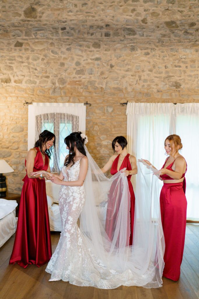 Bridesmaids in crimson dresses assisting with the bride's gown preparations.