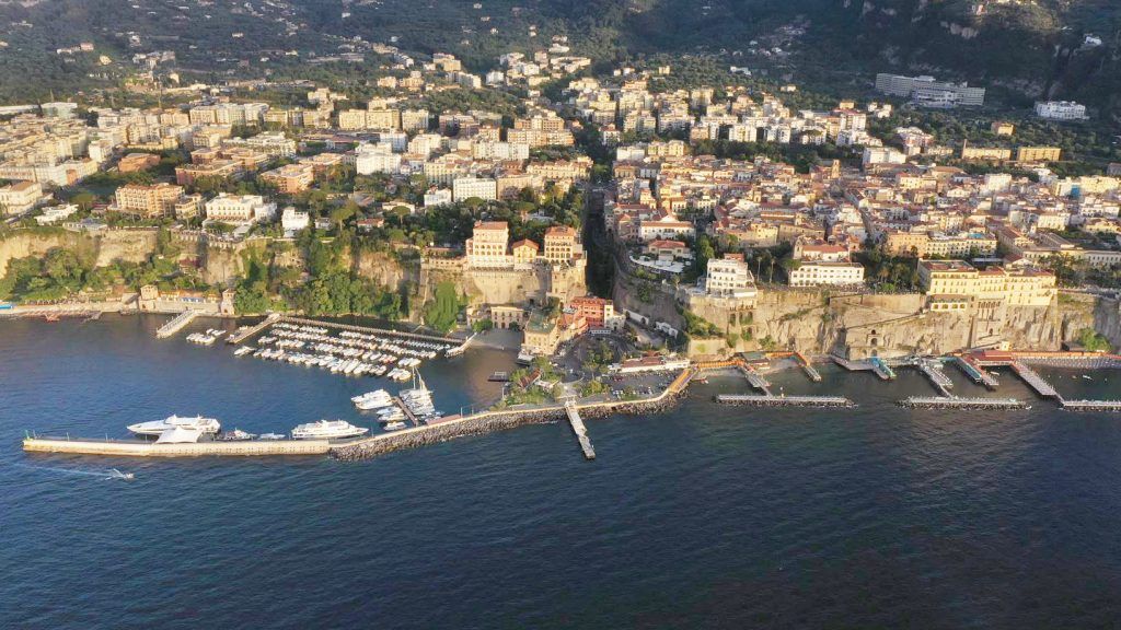 Aerial panoramic view of a coastal Italian town with mountains and harbor in the background.