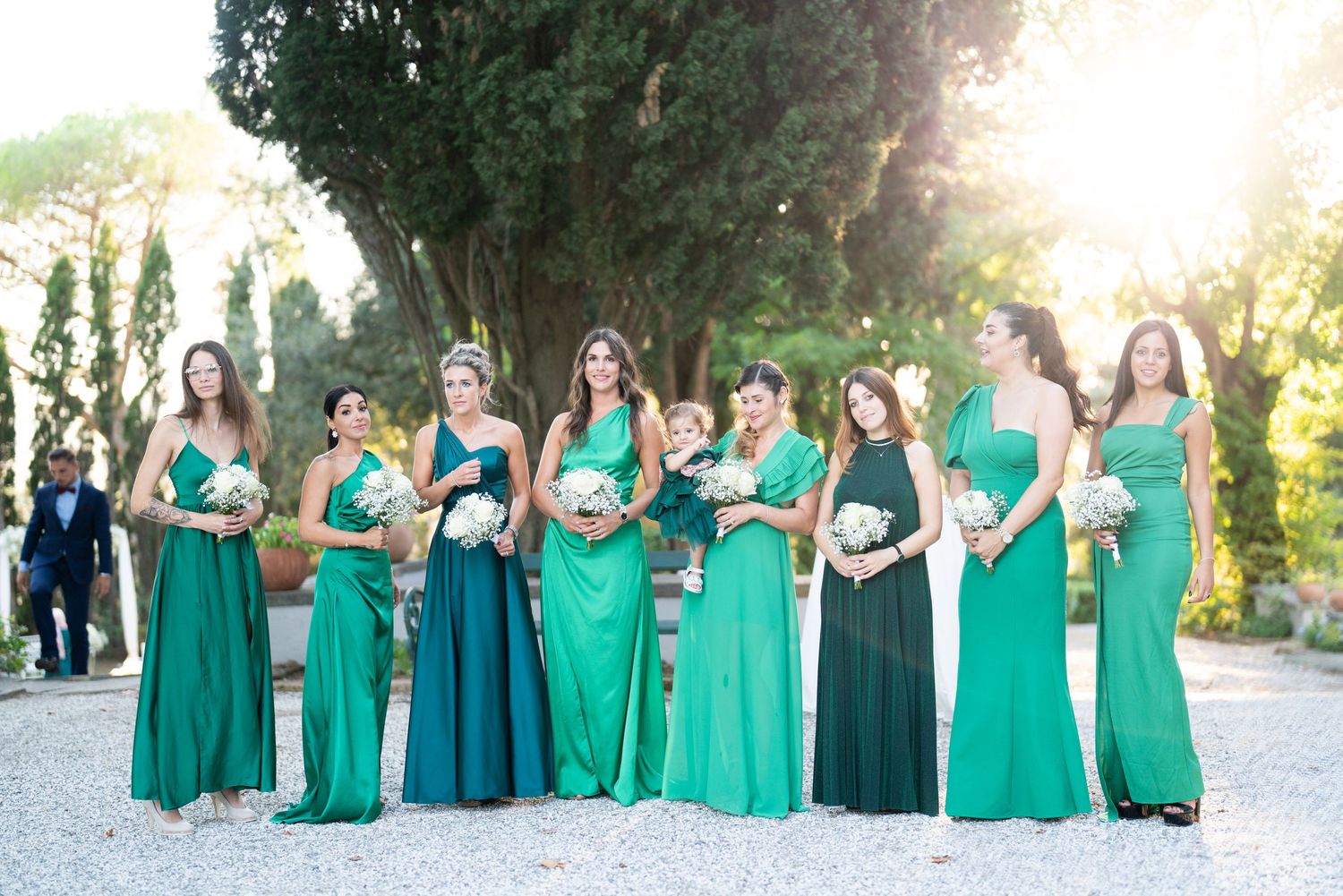 Bridesmaids in coordinating emerald green dresses pose together at an outdoor wedding venue.