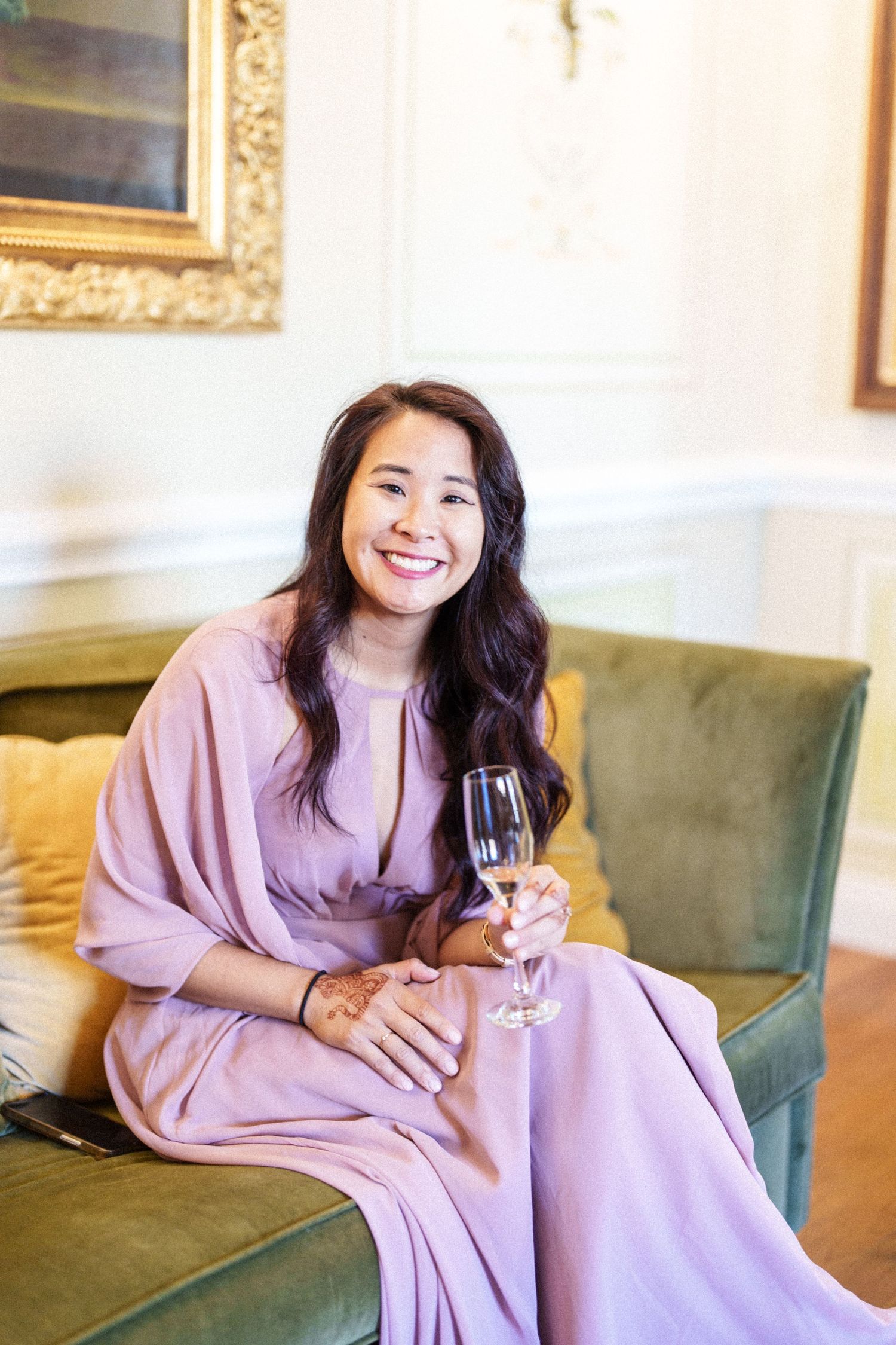 A woman in a pink robe relaxes on a green velvet couch with a champagne glass.