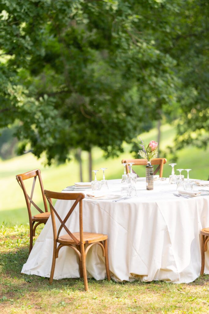 Elegant outdoor wedding reception setup with round tables covered in white linens and wooden cross-back chairs.
