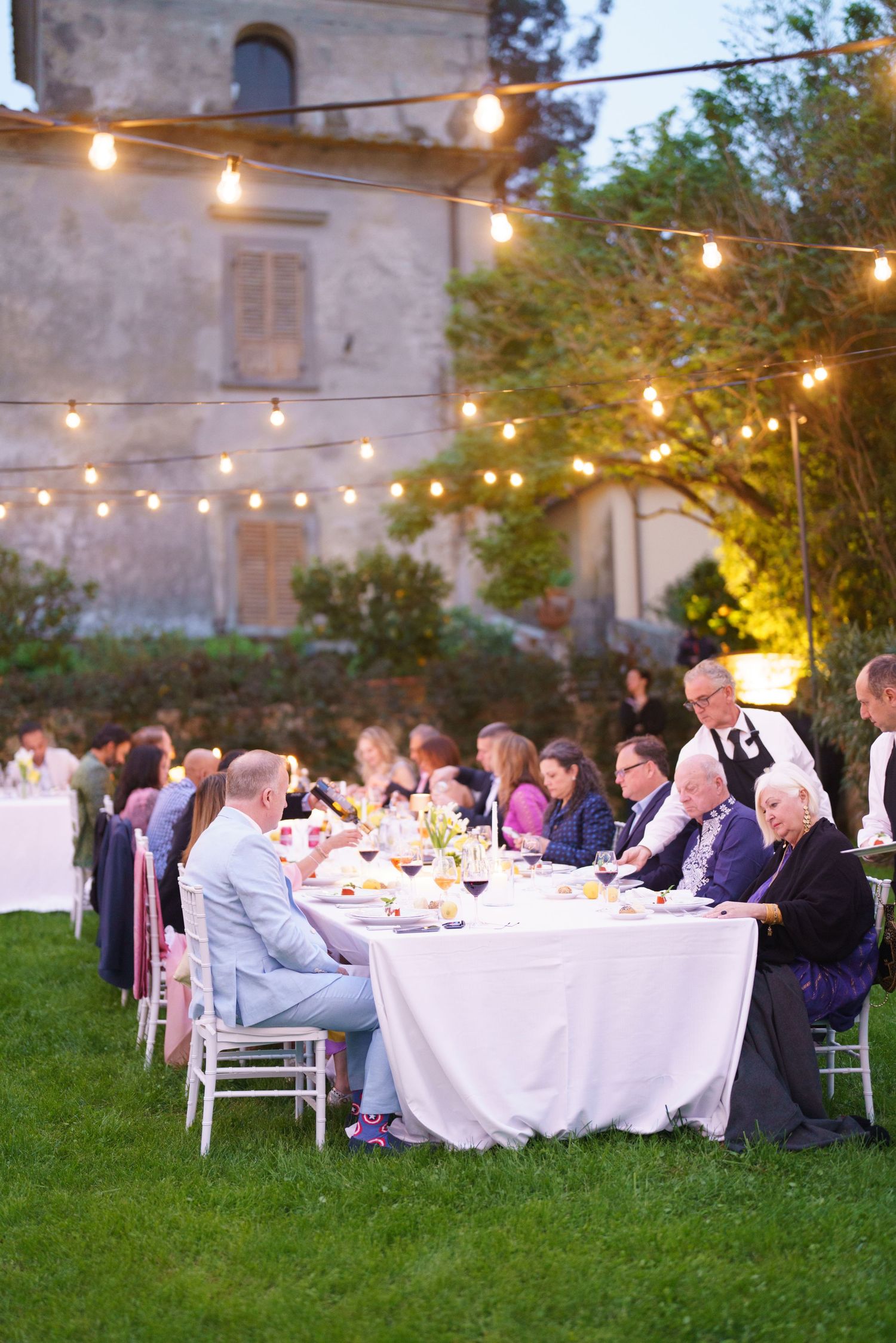 Evening wedding reception with guests dining at long tables under string lights.