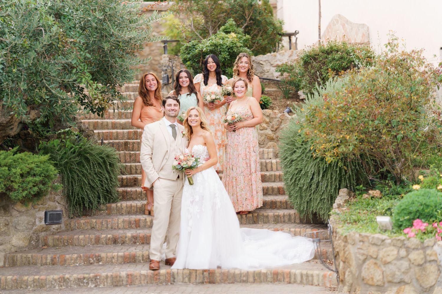 Bridal party members in soft pastel dresses pose on rustic stone steps.