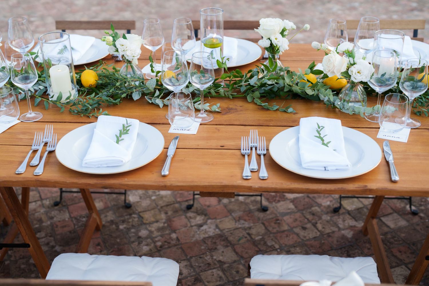 Elegant outdoor wedding table setting with white plates, linen napkins, greenery garland, yellow flowers, and crystal glassware.
