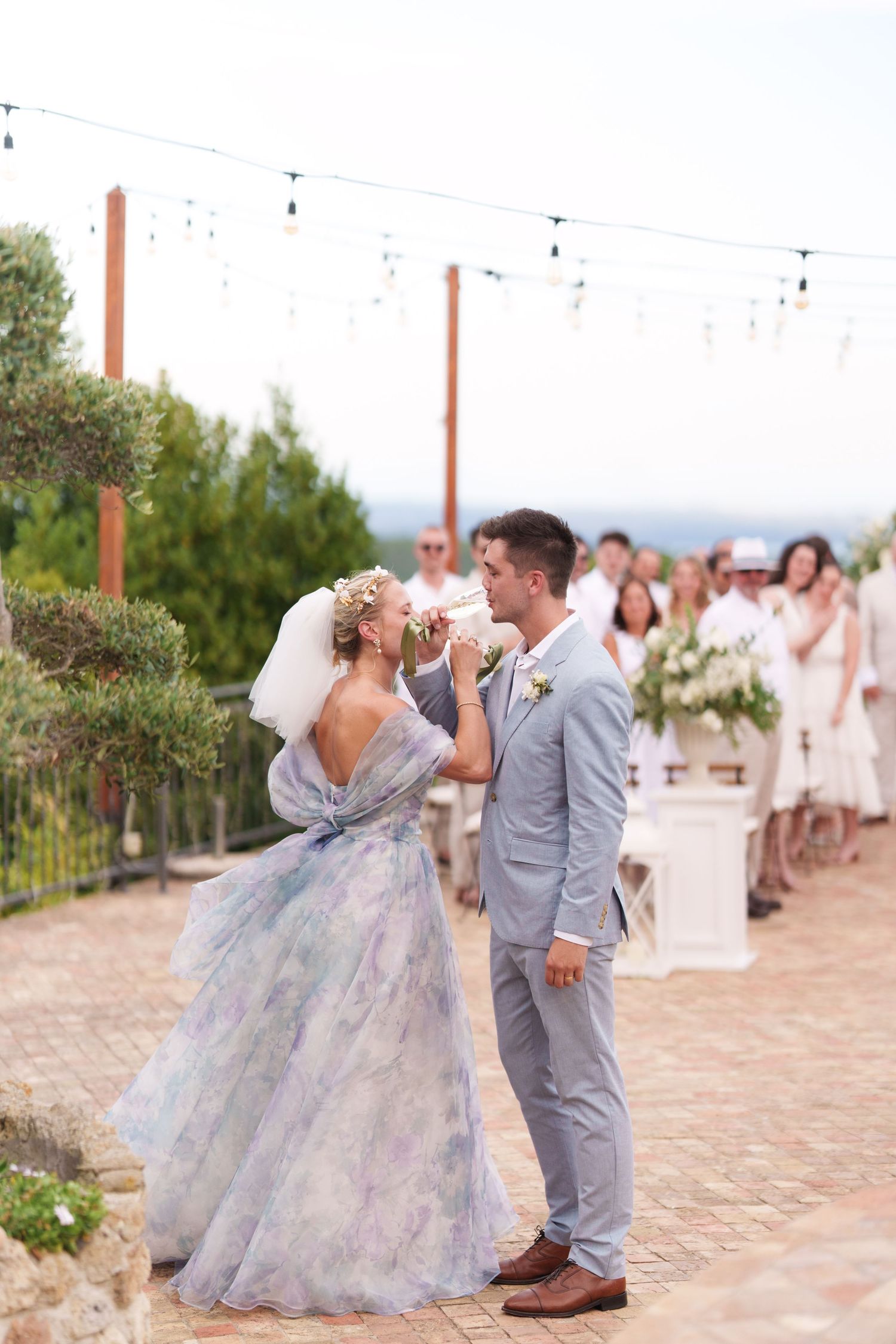 Bride in floral gown and groom in gray suit share a kiss during outdoor wedding ceremony with string lights.