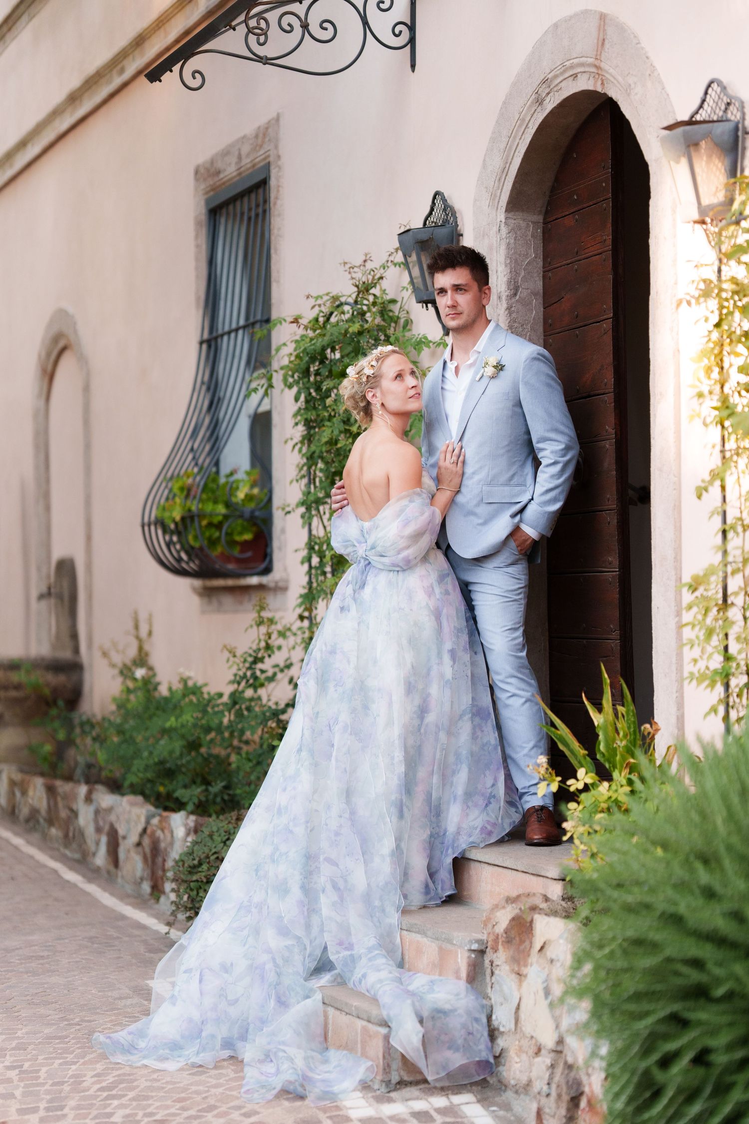 Bride in blue floral gown and groom in light blue suit pose elegantly at a stone villa entrance surrounded by greenery.