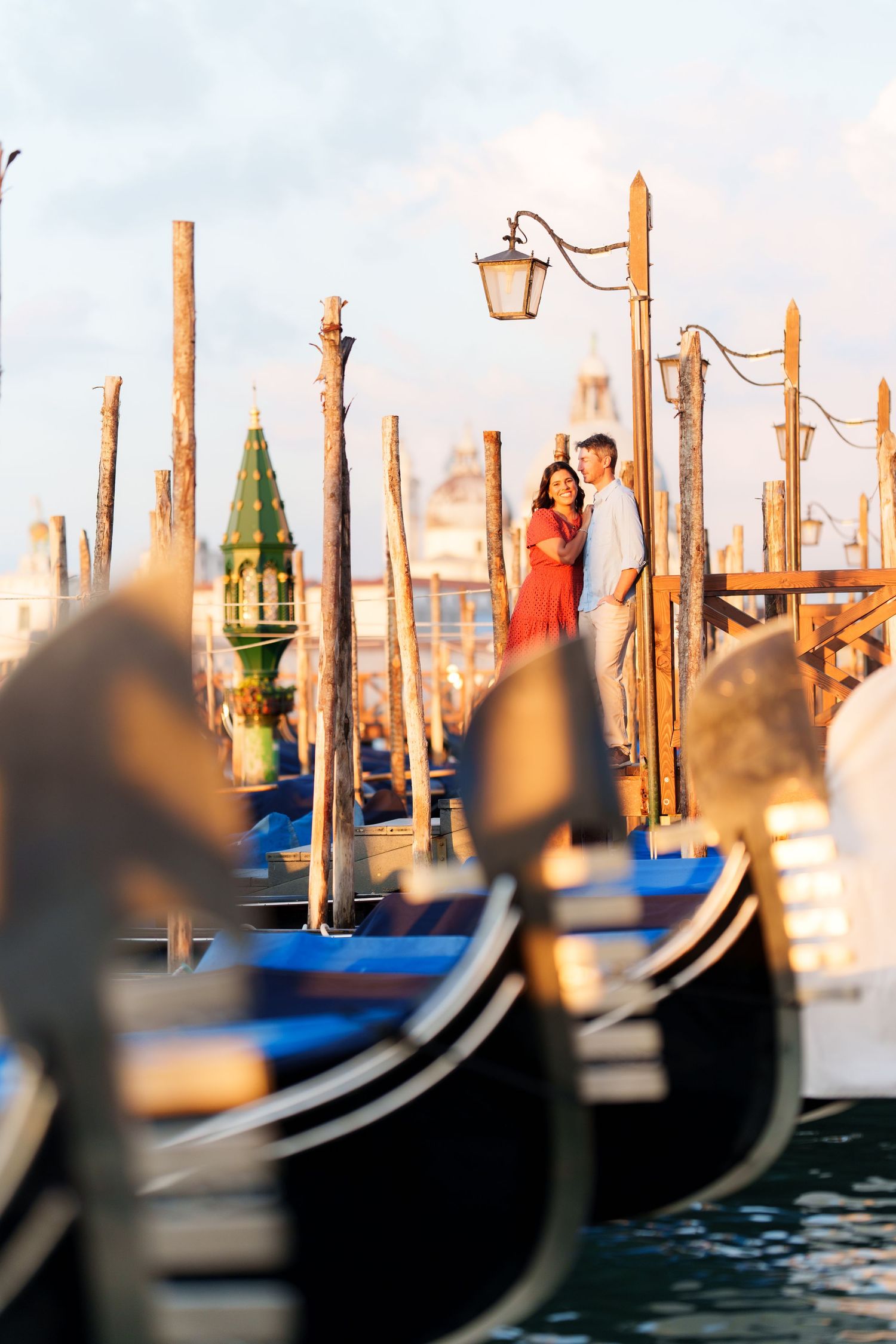 Romantic couple embracing on Venice gondola dock with classic wooden poles and Santa Maria della Salute in background.