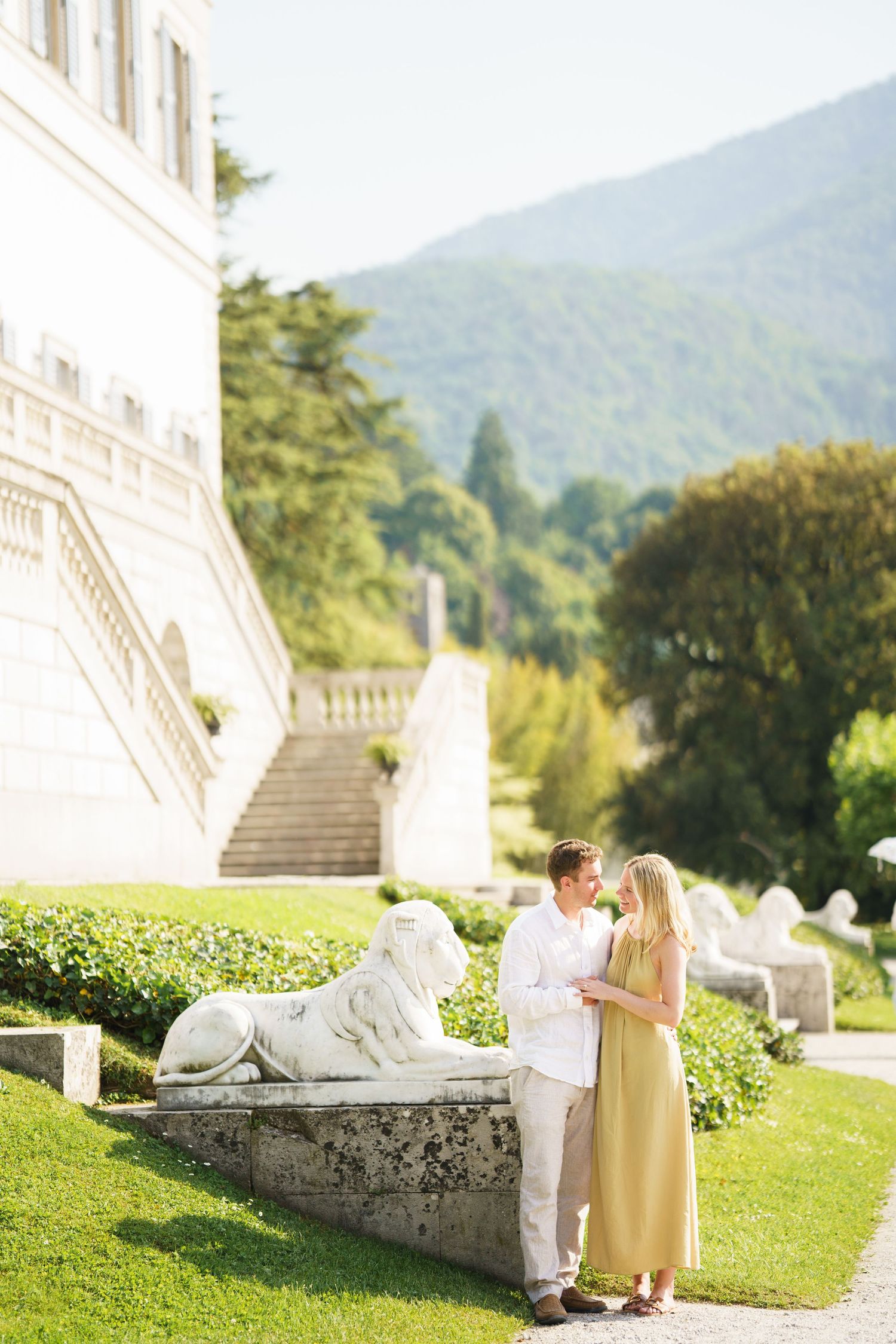 Couple standing in a sunlit garden near a white mansion with lion statues, green lawn, and mountain backdrop.