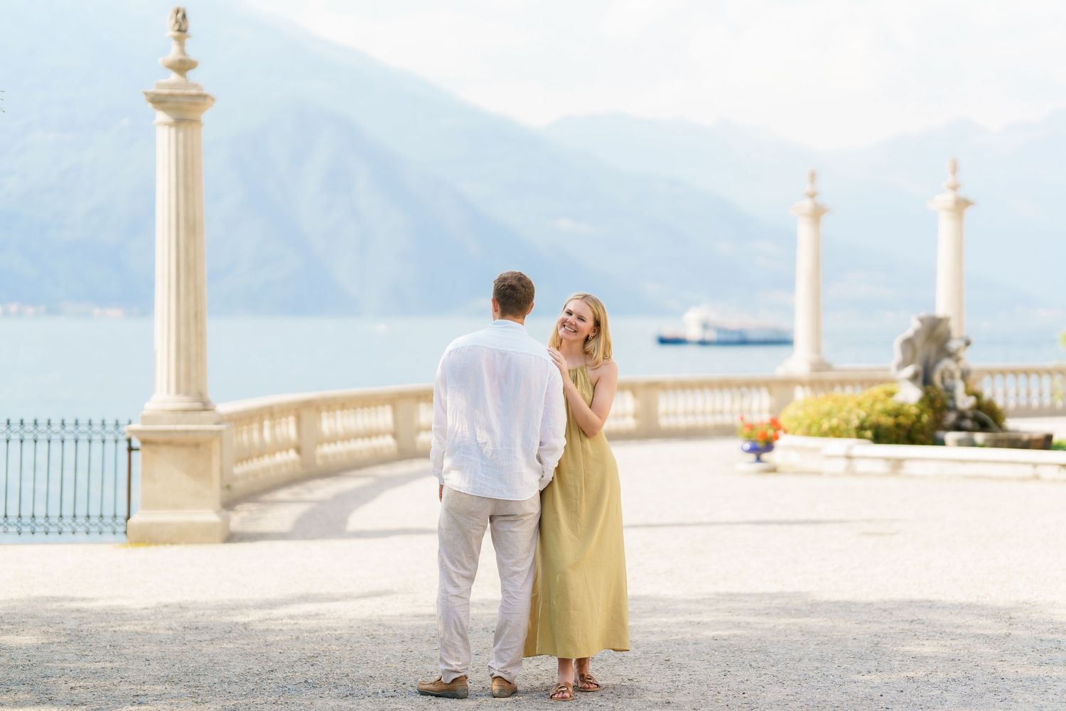 Couple holding hands on elegant lakeside terrace with mountain backdrop and ornate columns.