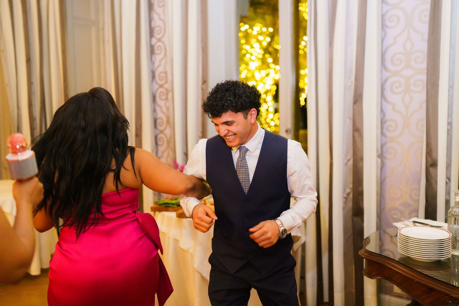 Man in navy vest dancing with woman in pink dress at festive indoor reception event.