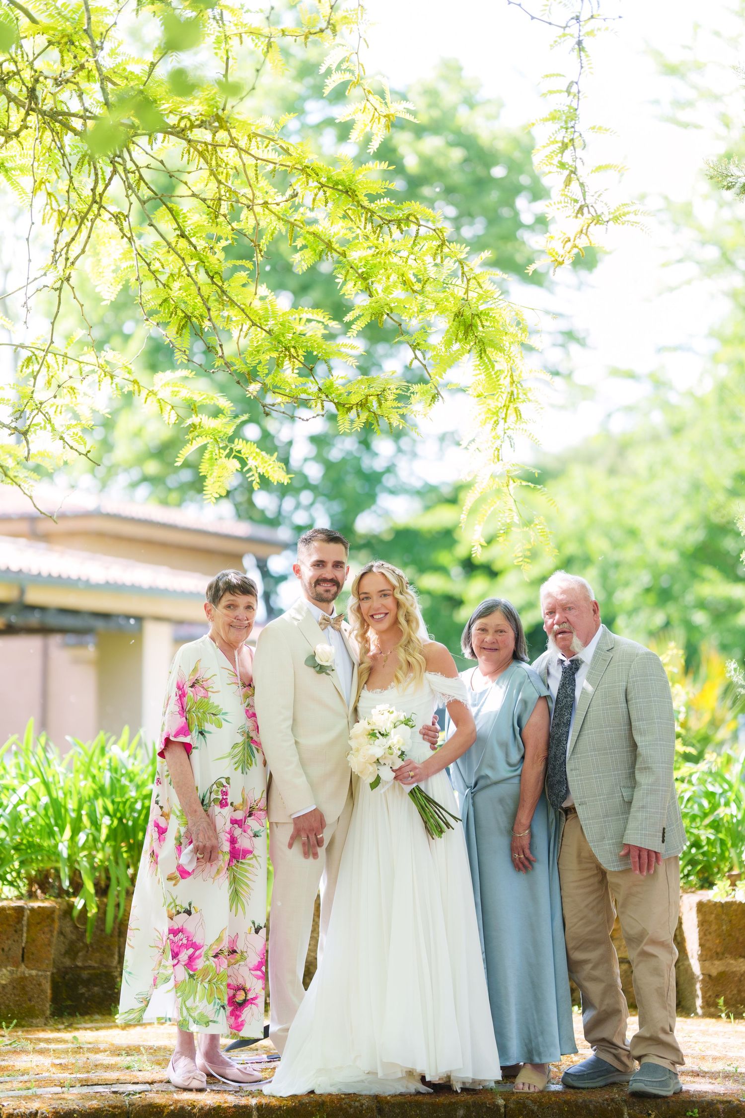 Bride and groom pose with family outdoors in lush garden setting, bride holding white bouquet in elegant wedding dress.