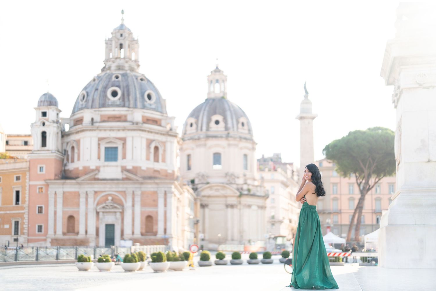 Woman in green gown poses before Rome's iconic twin domes and Trajan's Column in soft morning light.