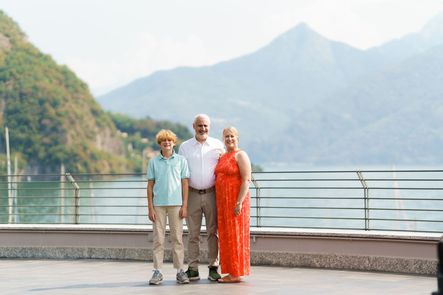 Family of three posing on lakeside promenade with mountain backdrop on sunny day.