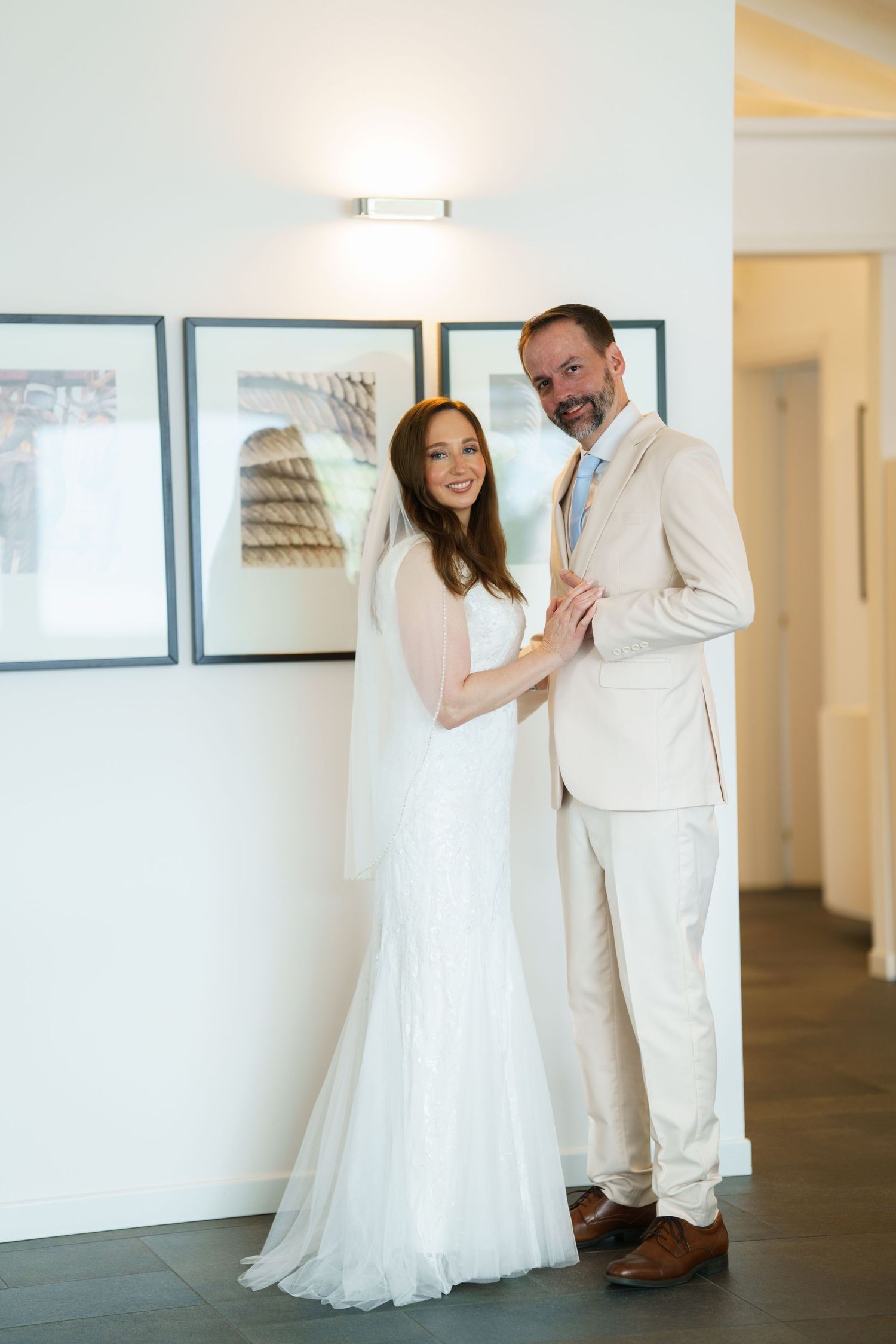 Bride in white gown and groom in beige suit smiling together indoors at their wedding venue.