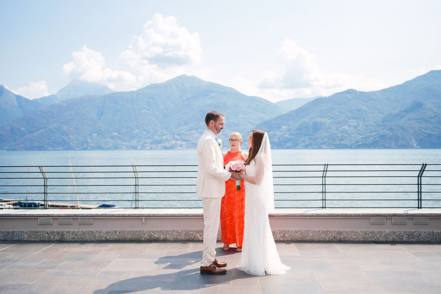 Couple exchanges vows with officiant on lakeside terrace with mountain backdrop on their wedding day.