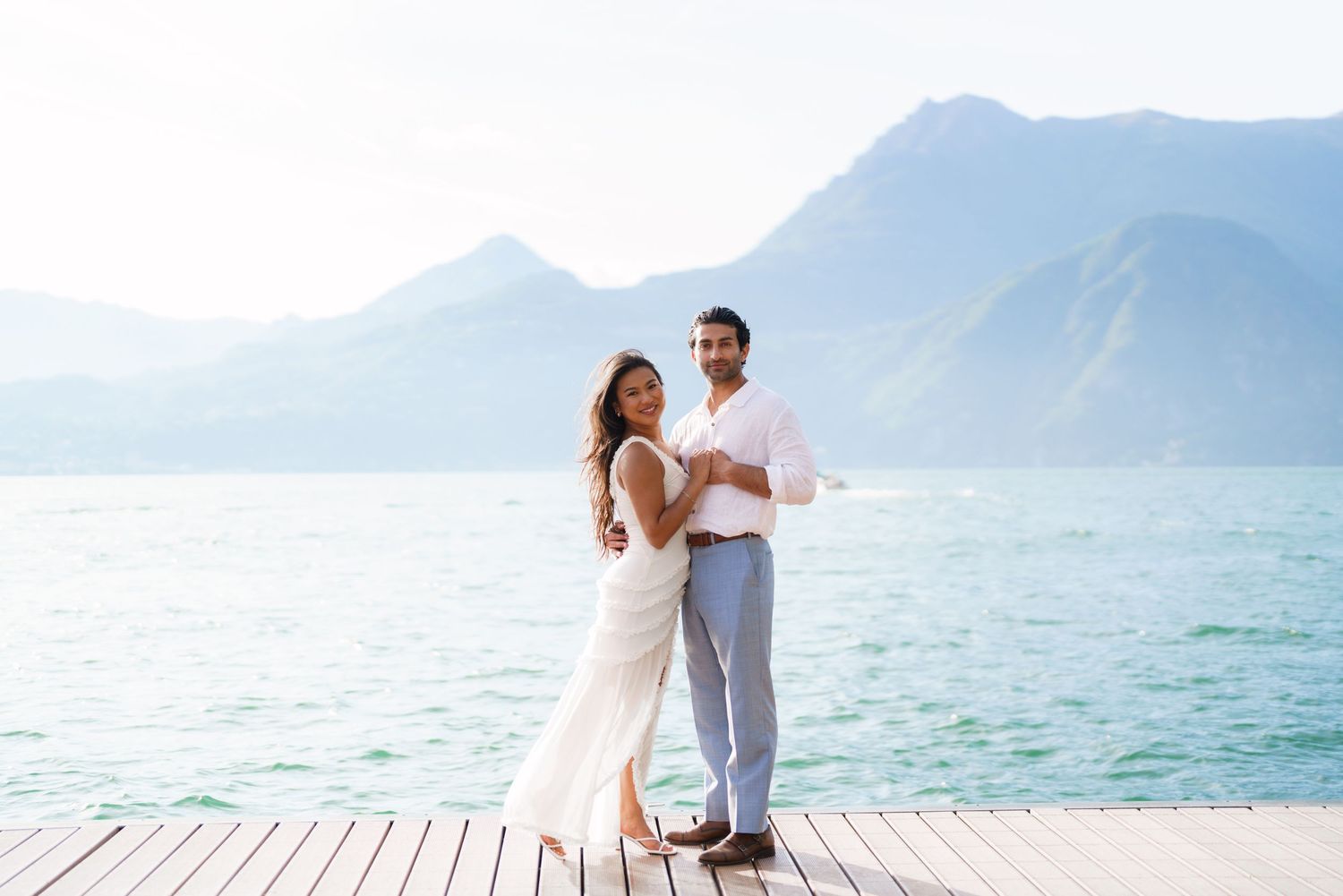 Couple in white outfits posing on a wooden dock with turquoise lake and mountains in the background.